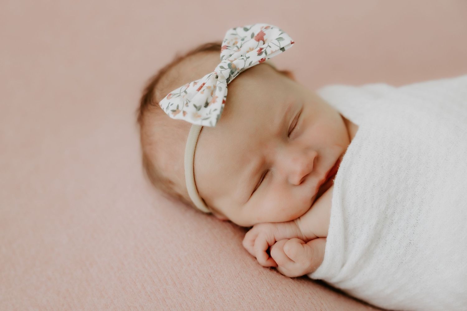 Peaceful sleeping newborn wearing a delicate lace bow headband on pink background.
