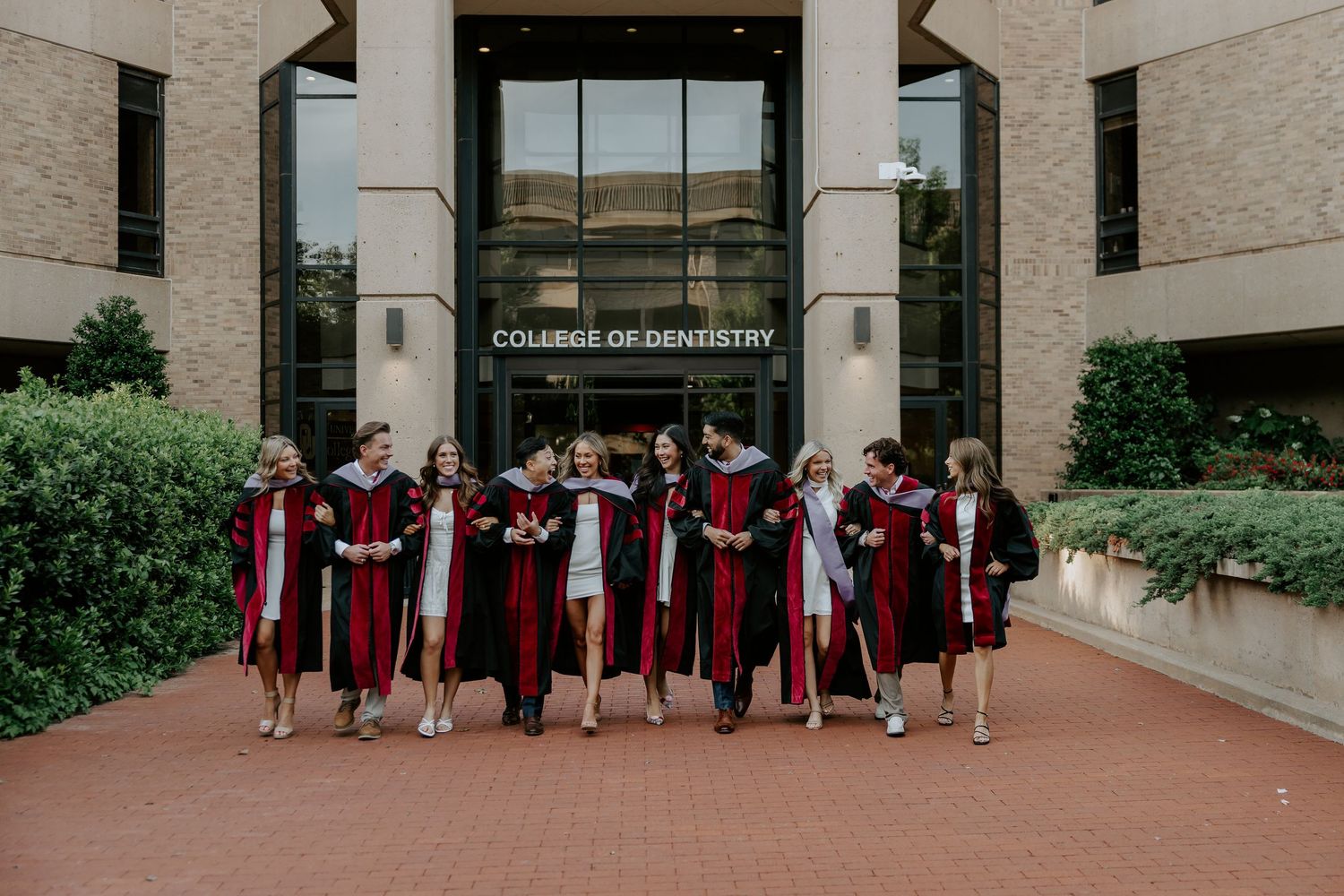Group of graduates in red and black robes walking together outside a college building.