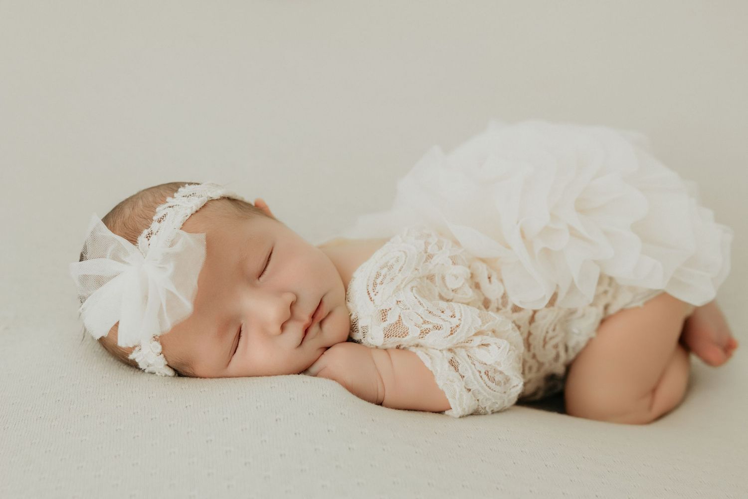 Peaceful sleeping newborn wrapped in delicate white lace fabric against neutral background.