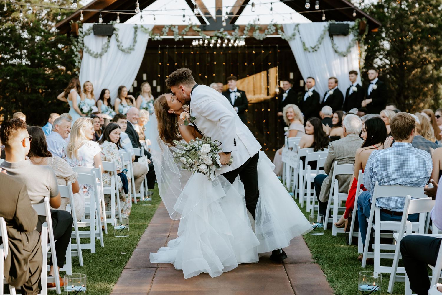 A couple shares their first kiss as newlyweds during an outdoor garden ceremony with white chairs.