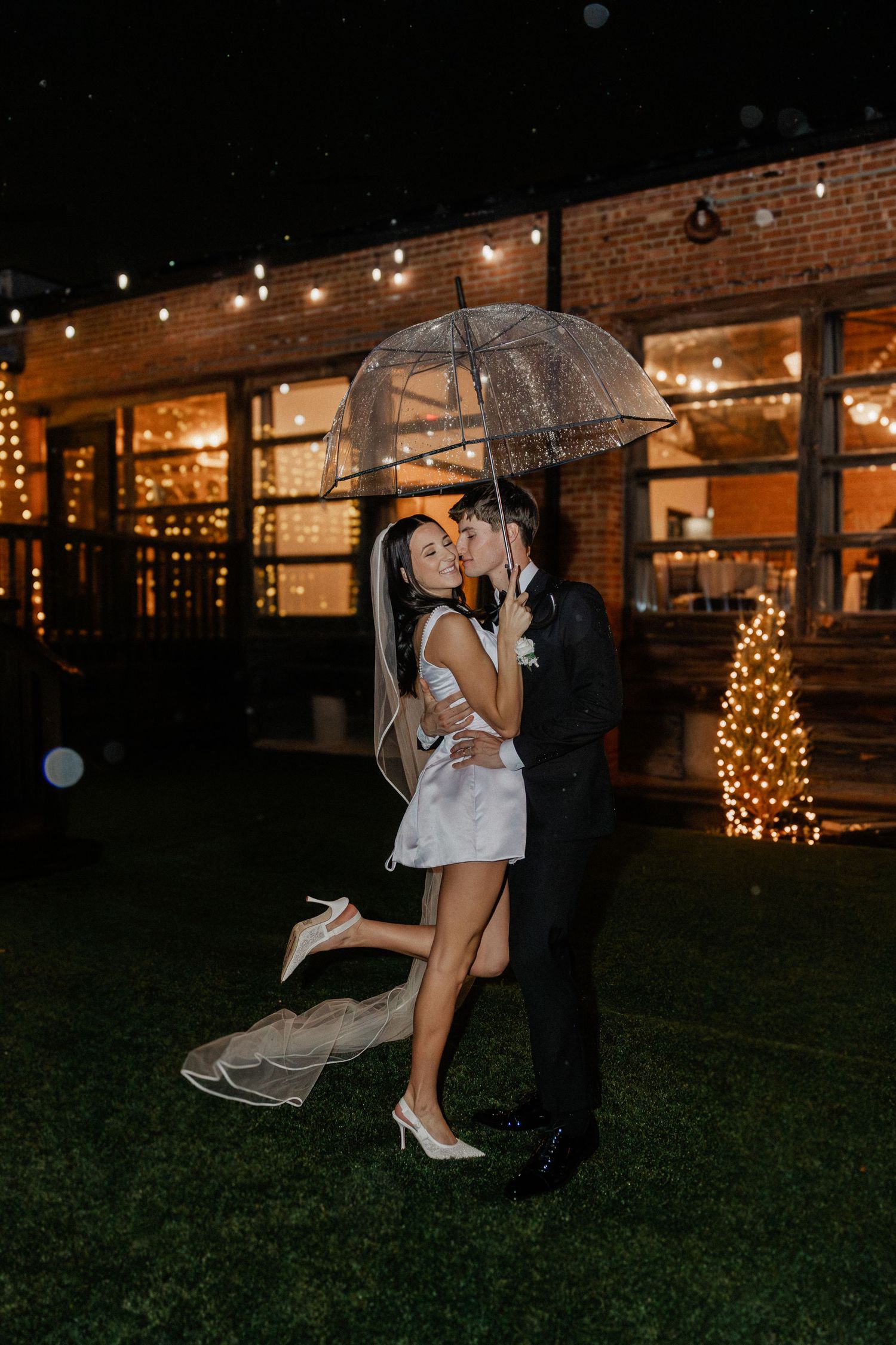 A romantic couple shares a kiss under a clear umbrella at night with twinkling lights in the background.