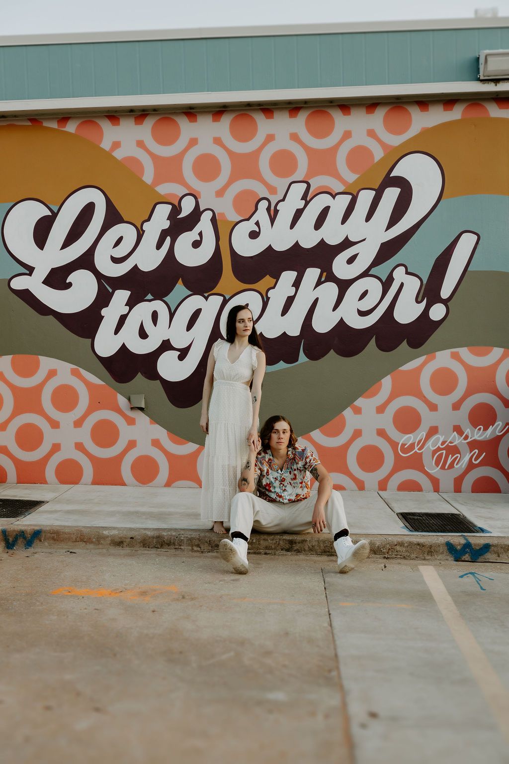 A couple poses playfully in front of a colorful wall mural with retro typography.
