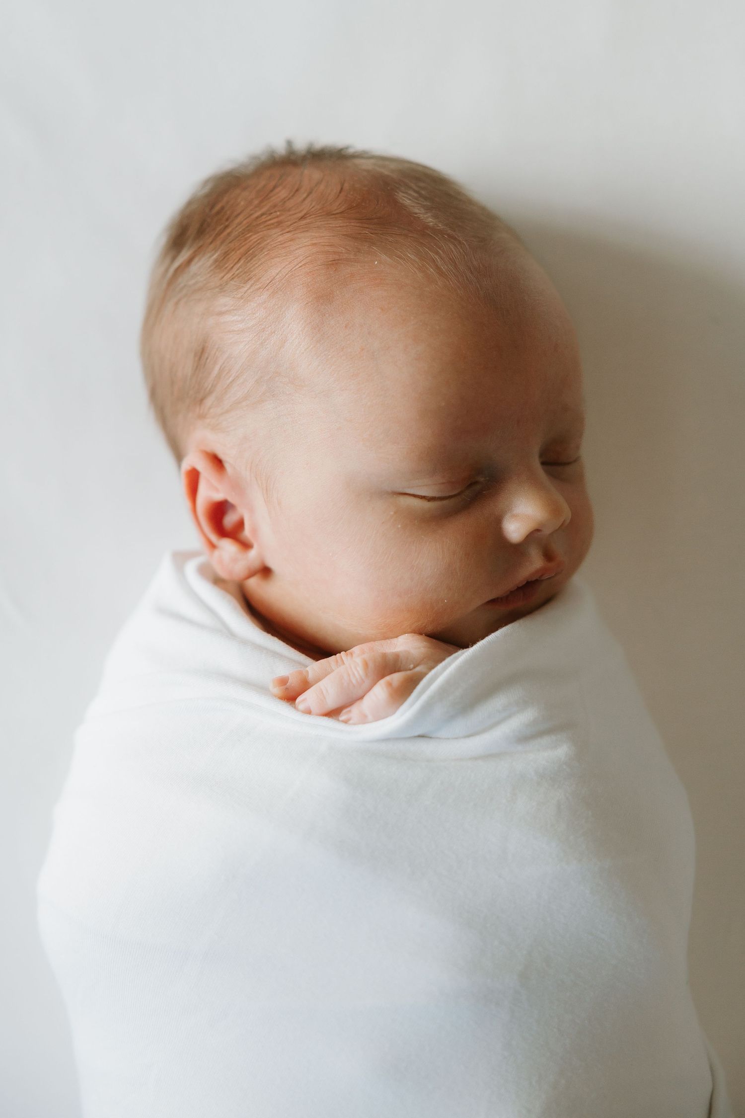 Newborn baby swaddled snugly in soft white wrap against light background.