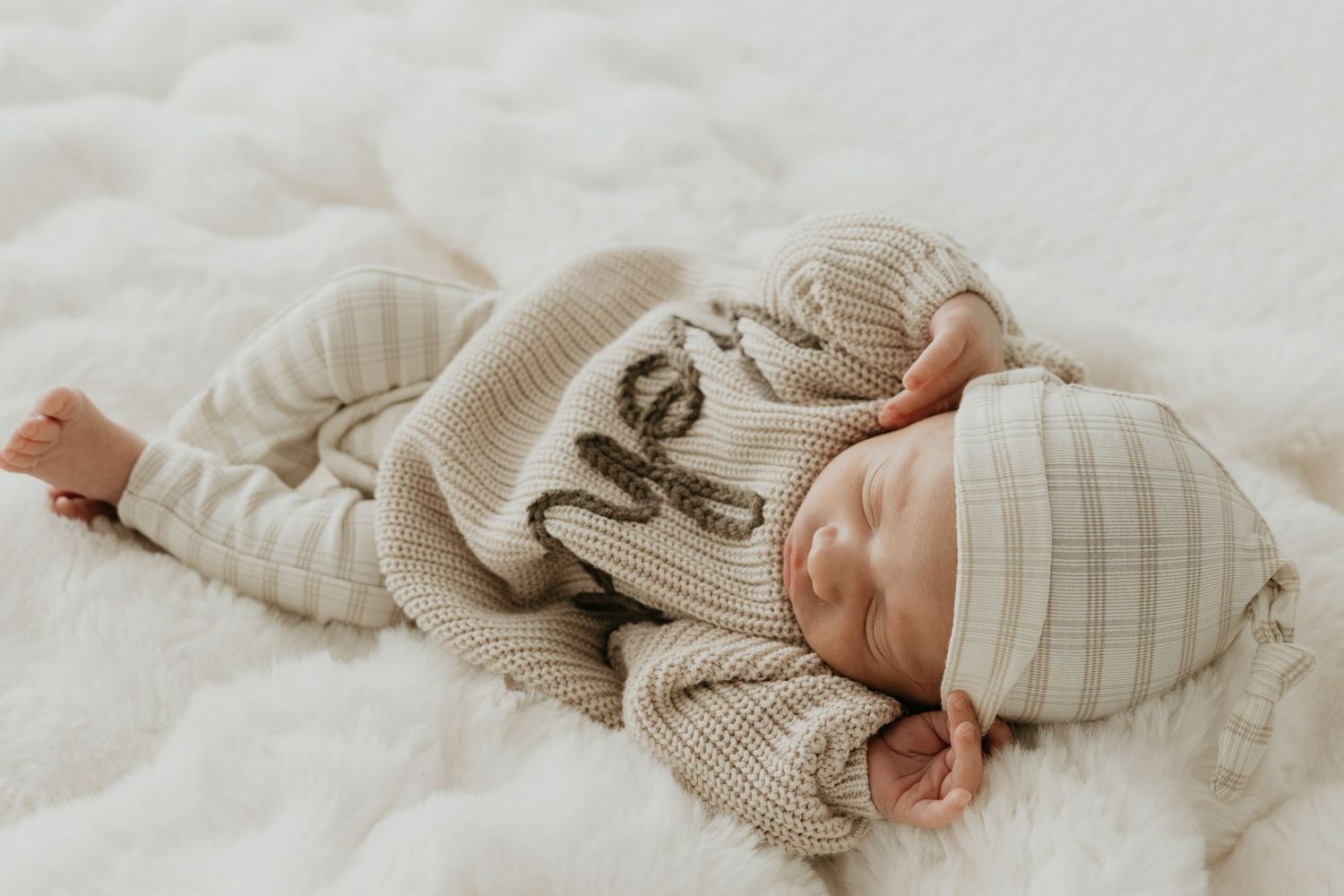 Sleeping infant wearing neutral colored knitwear on white bedding.