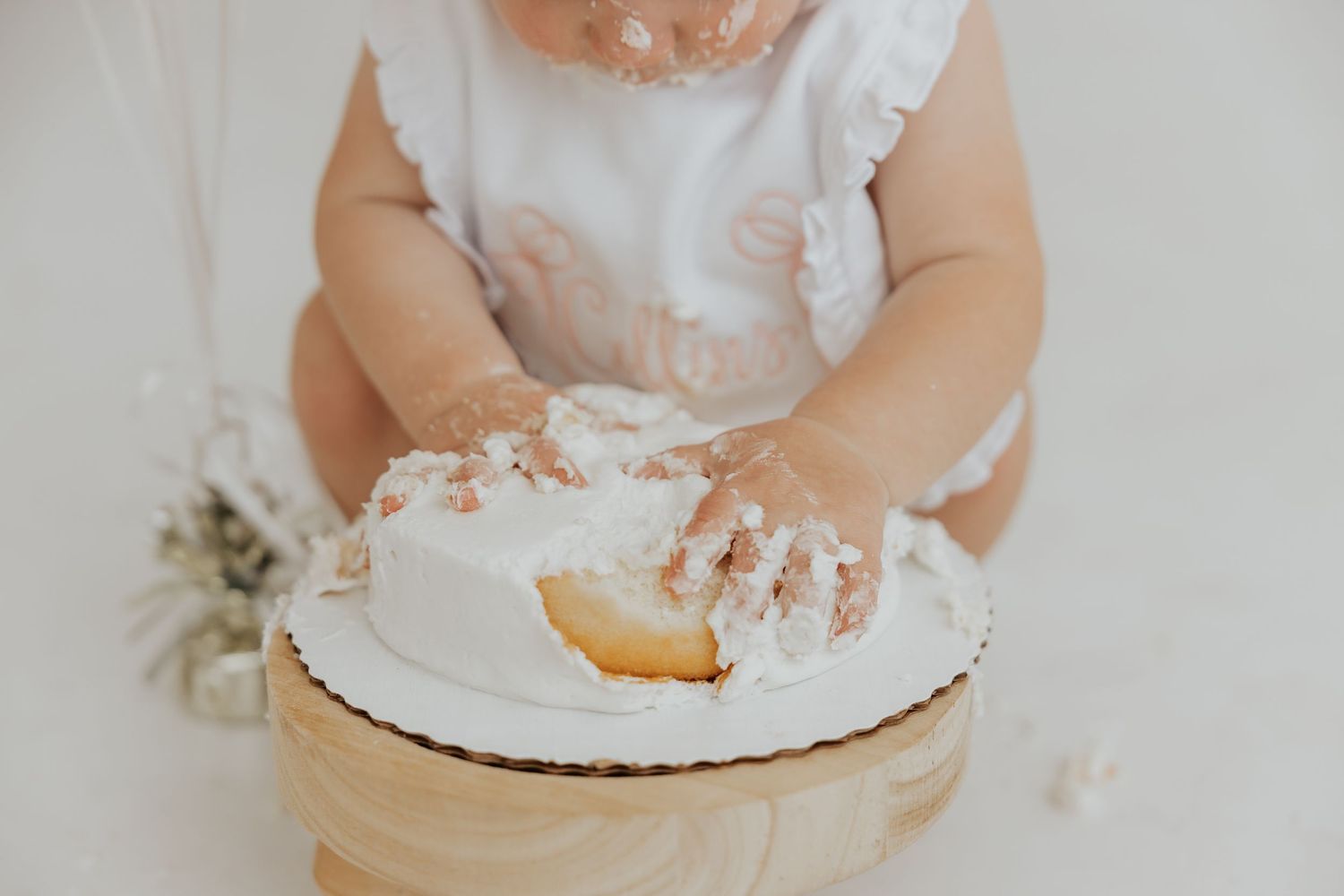 Baby in white outfit plays with cake and frosting on white background.