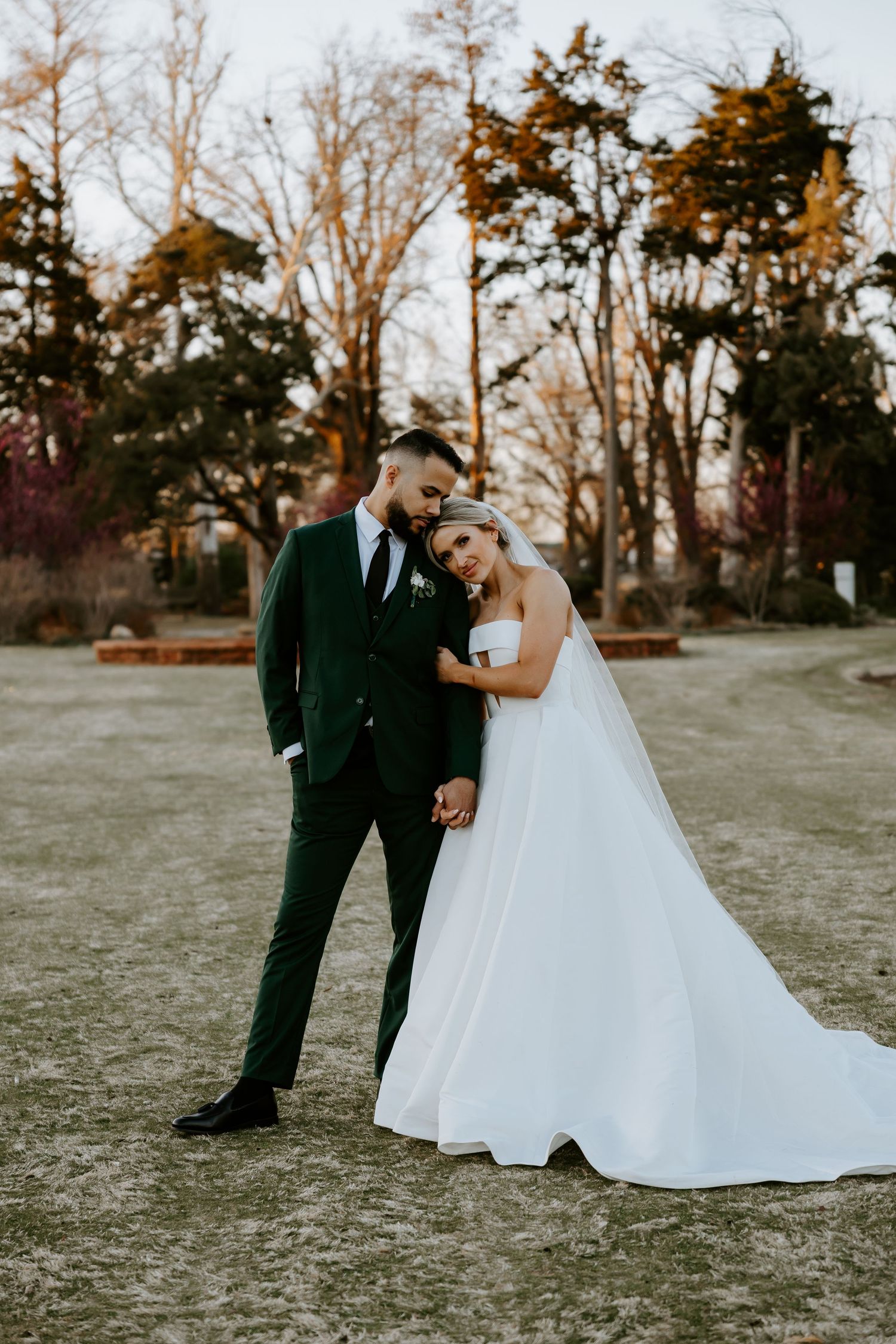 Wedding couple poses in an outdoor setting with bare trees and natural lighting at sunset.