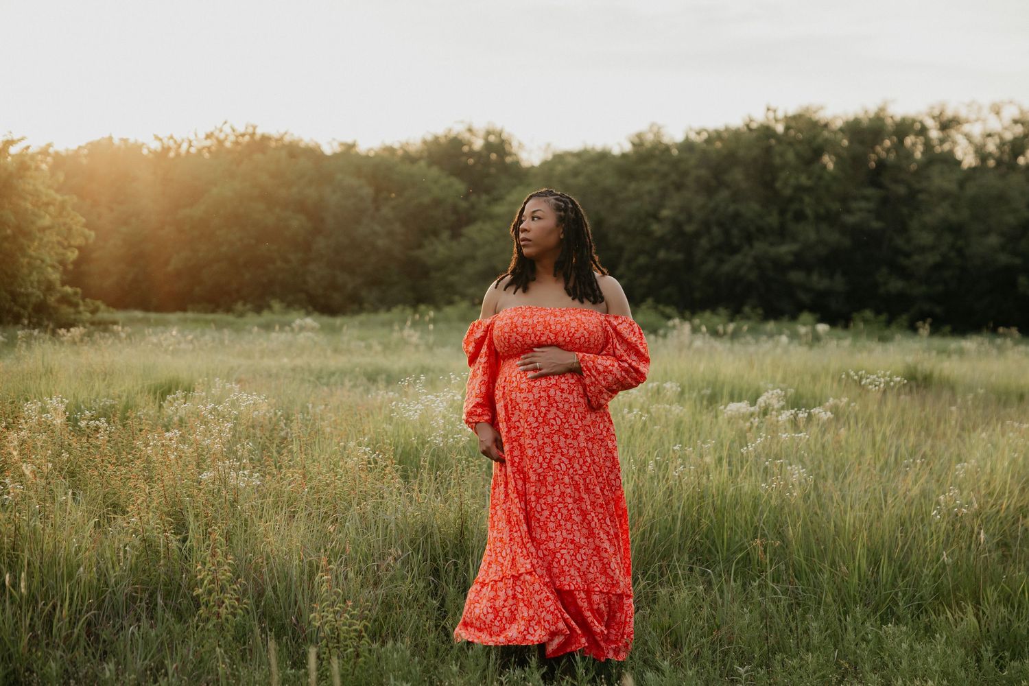 A flowing red floral maternity dress catches the evening light in a summer meadow.