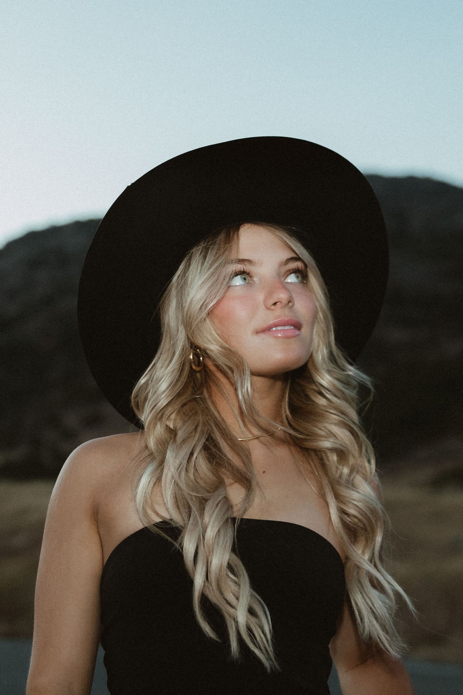 Stylish portrait of someone wearing a wide-brimmed black hat against a mountain backdrop.