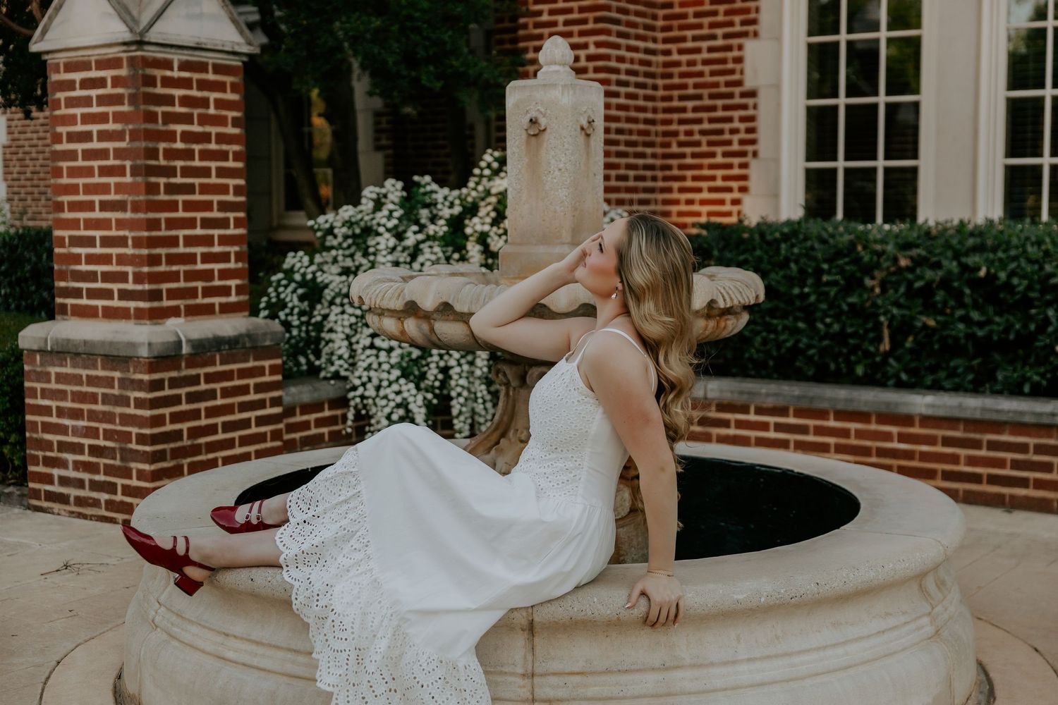 Graduate in white dress poses elegantly near brick building with windows.