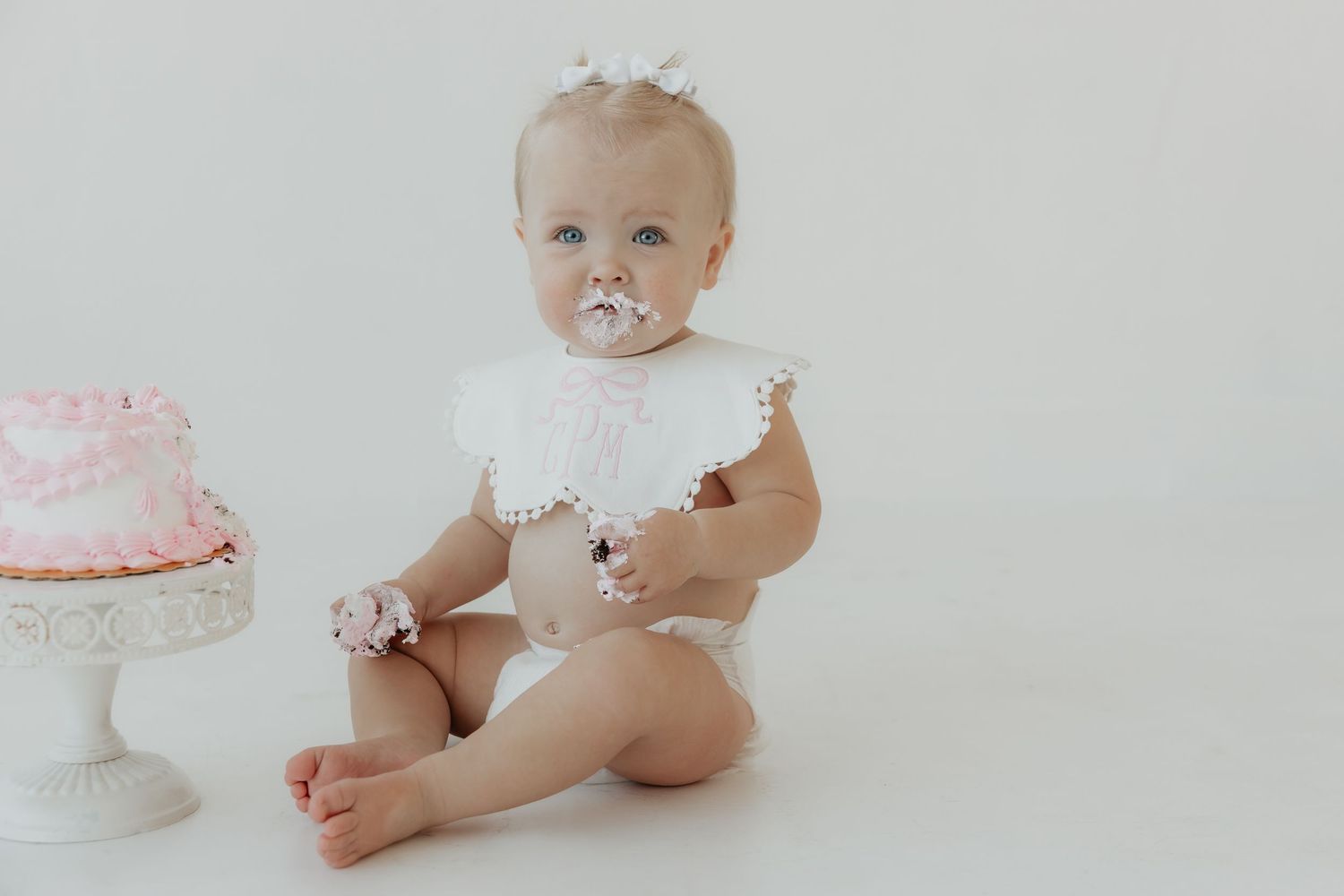 Pink and white first birthday cake smash photo session with delicate decorations and soft lighting.