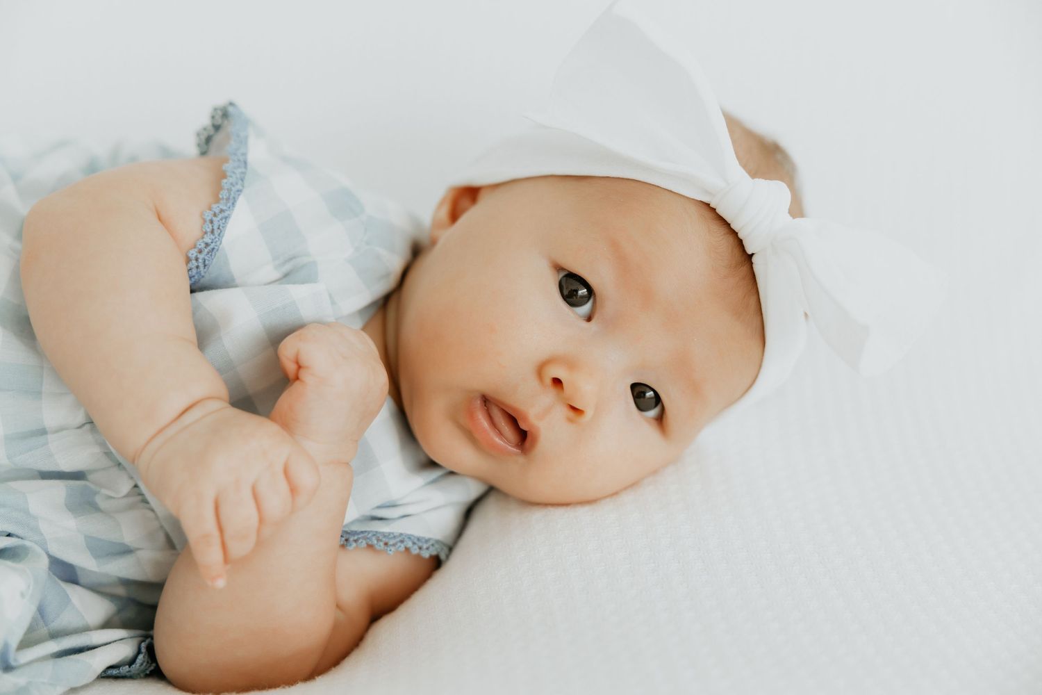 Baby wearing soft blue outfit and white hat resting on light bedding.