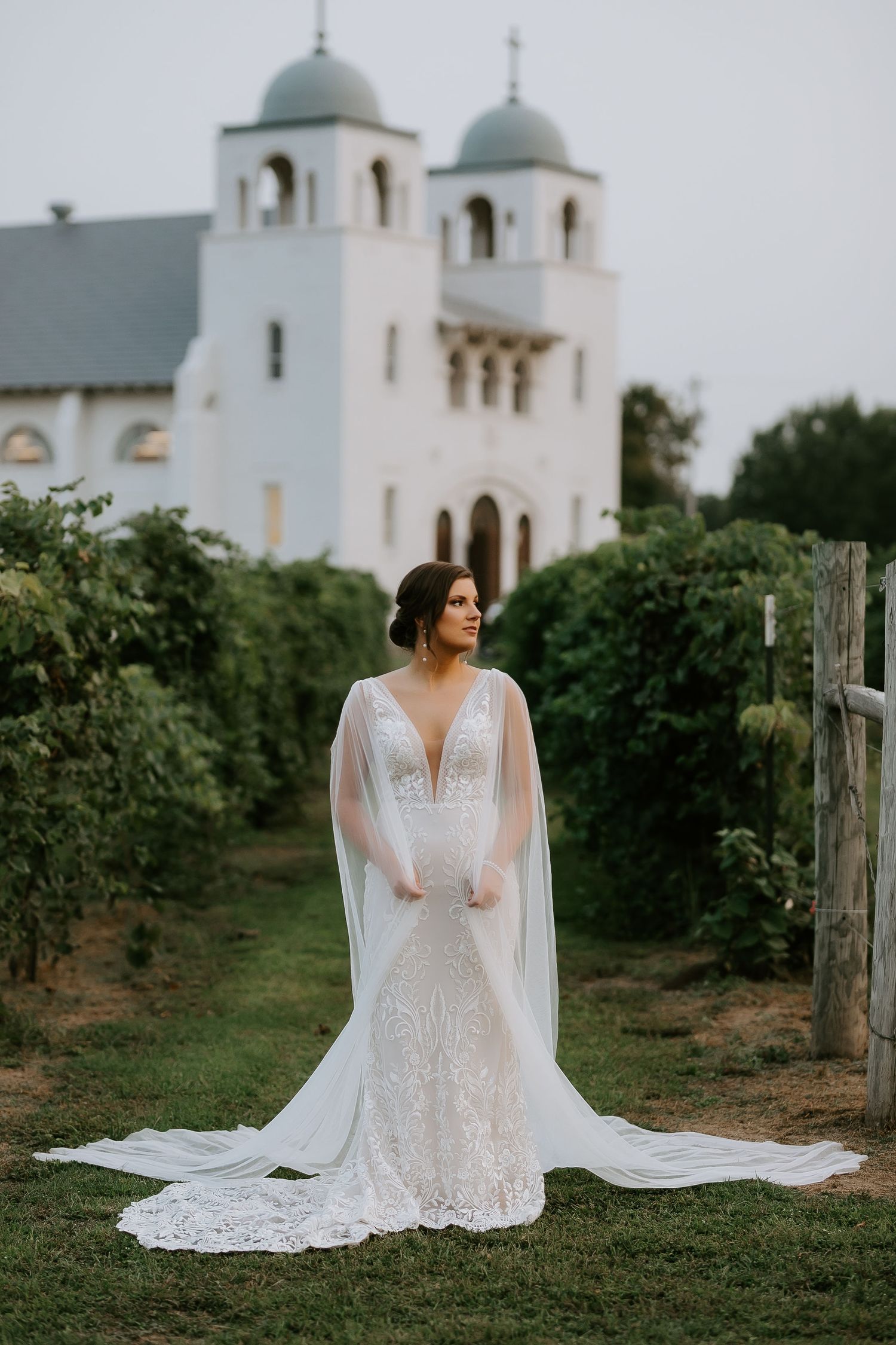 Bride in lace gown with cathedral veil poses near white church with domed towers.