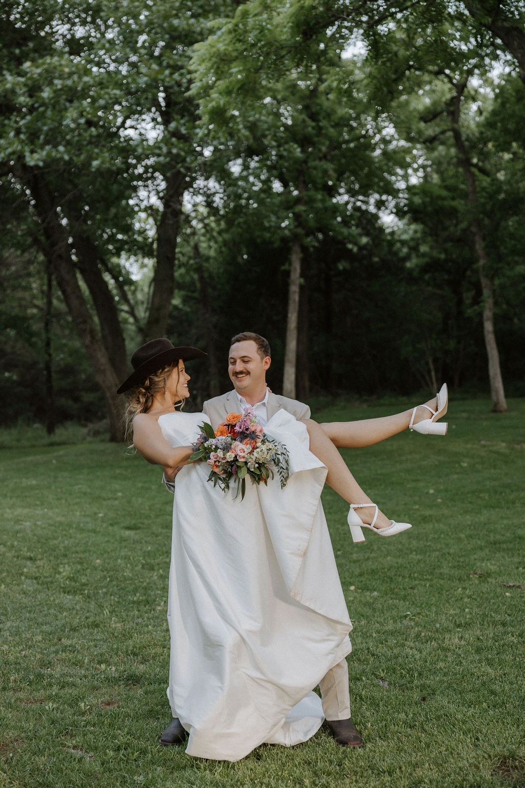 Playful wedding couple dances together on grassy lawn surrounded by trees.