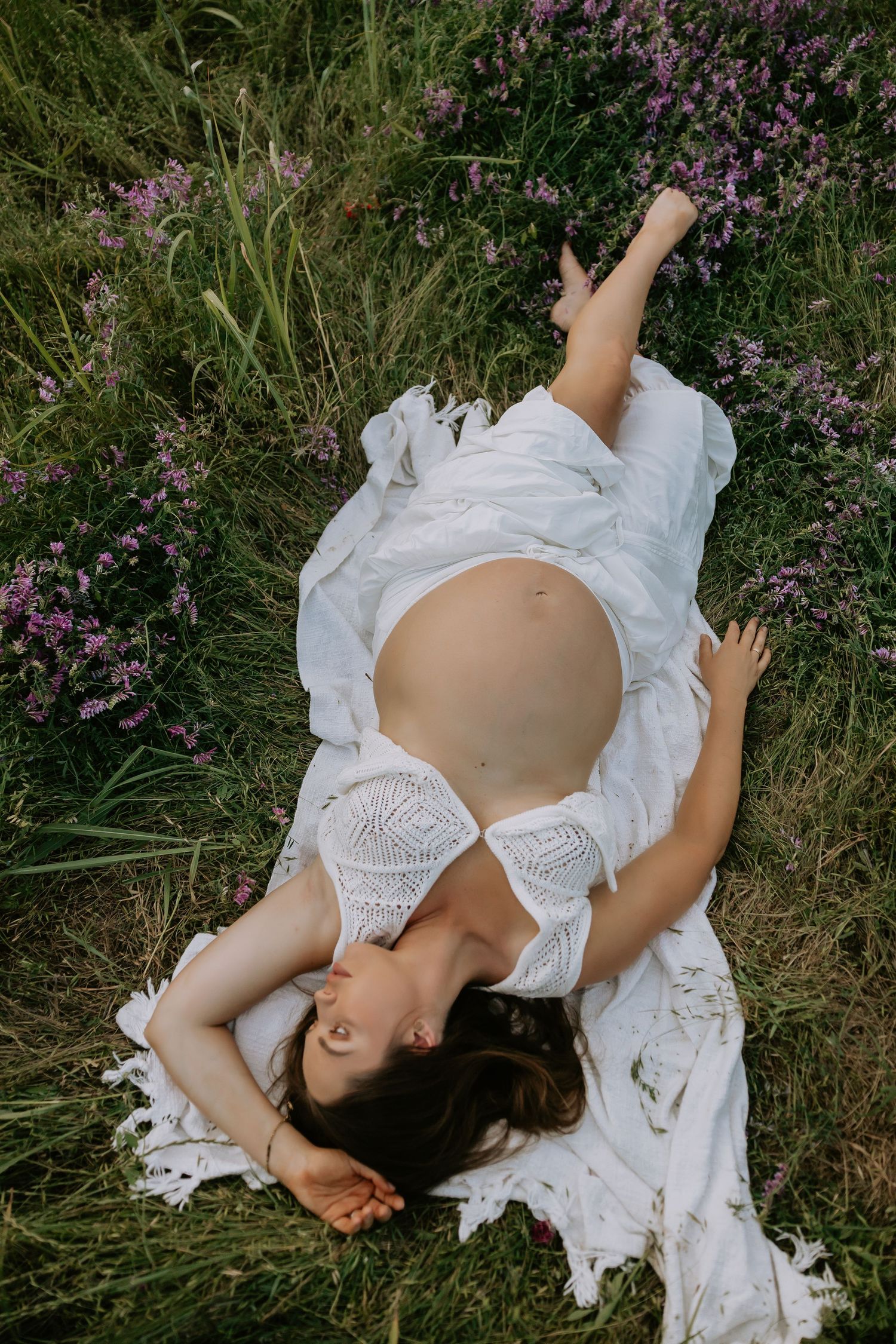 A dreamy maternity photo of someone lying in white lace among purple wildflowers.