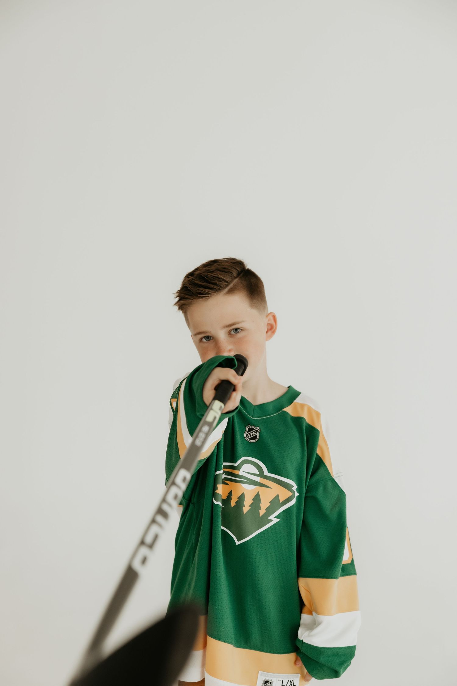 Young hockey fan poses with stick while wearing a Minnesota Wild NHL jersey against white background.