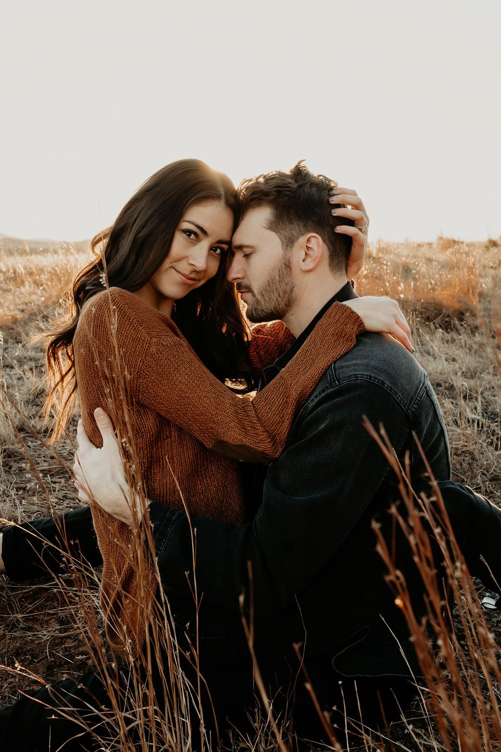 Intimate couple embracing in tall grass during golden hour, wearing brown and black outfits.