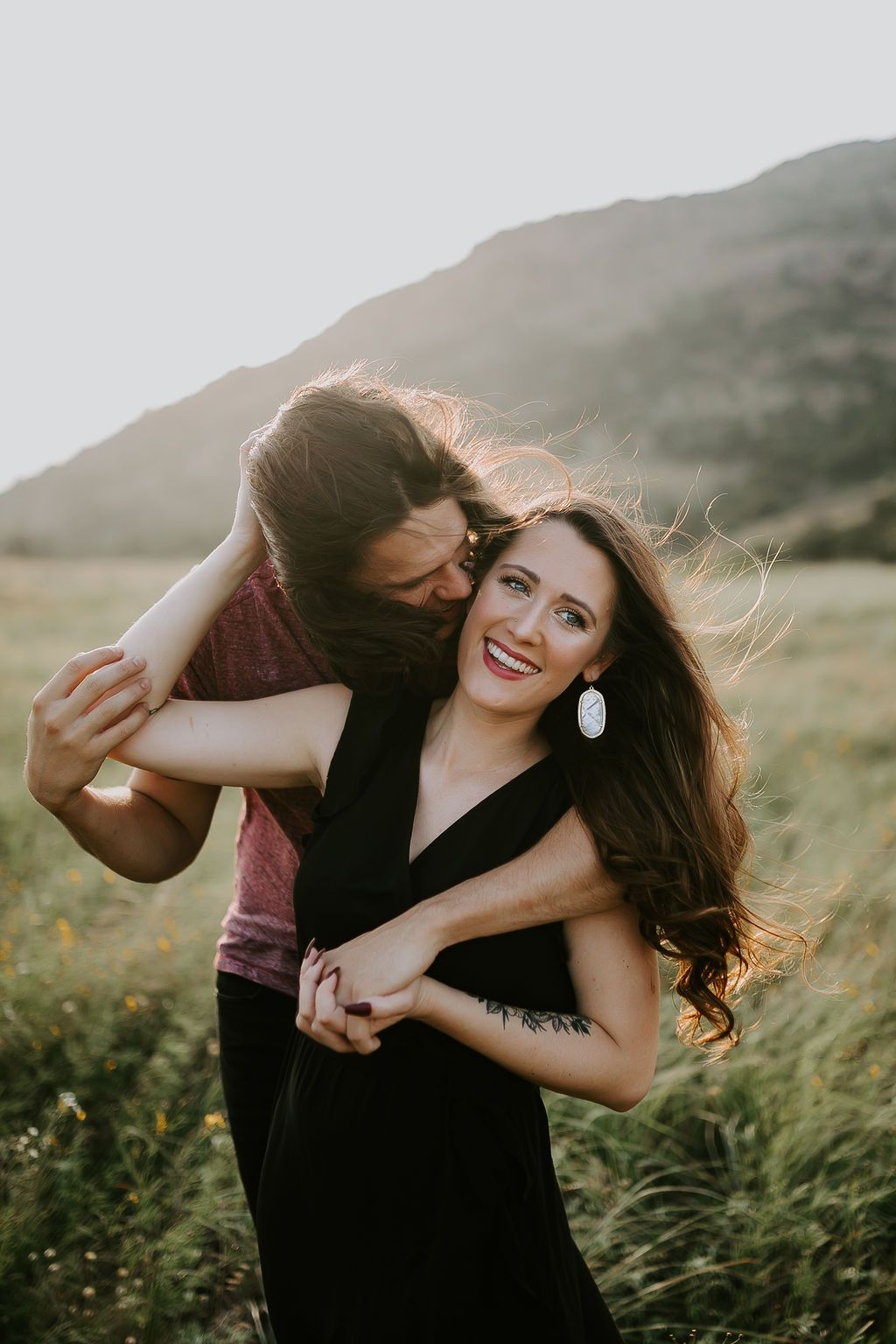 A romantic couple dances playfully in a field at sunset with tall grass and mountains in the background.