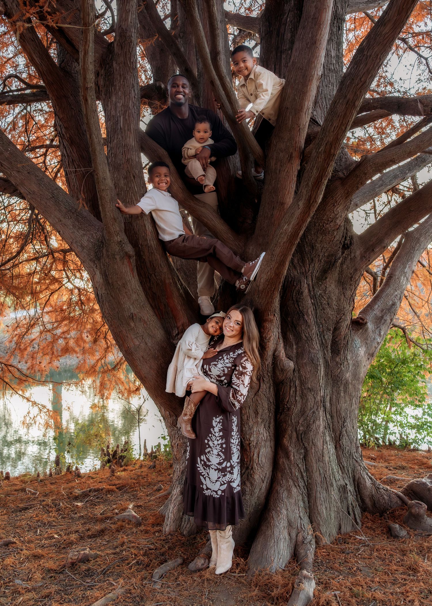 Family posing together in a large autumn tree near a lake, wearing coordinated outfits in fall colors.
