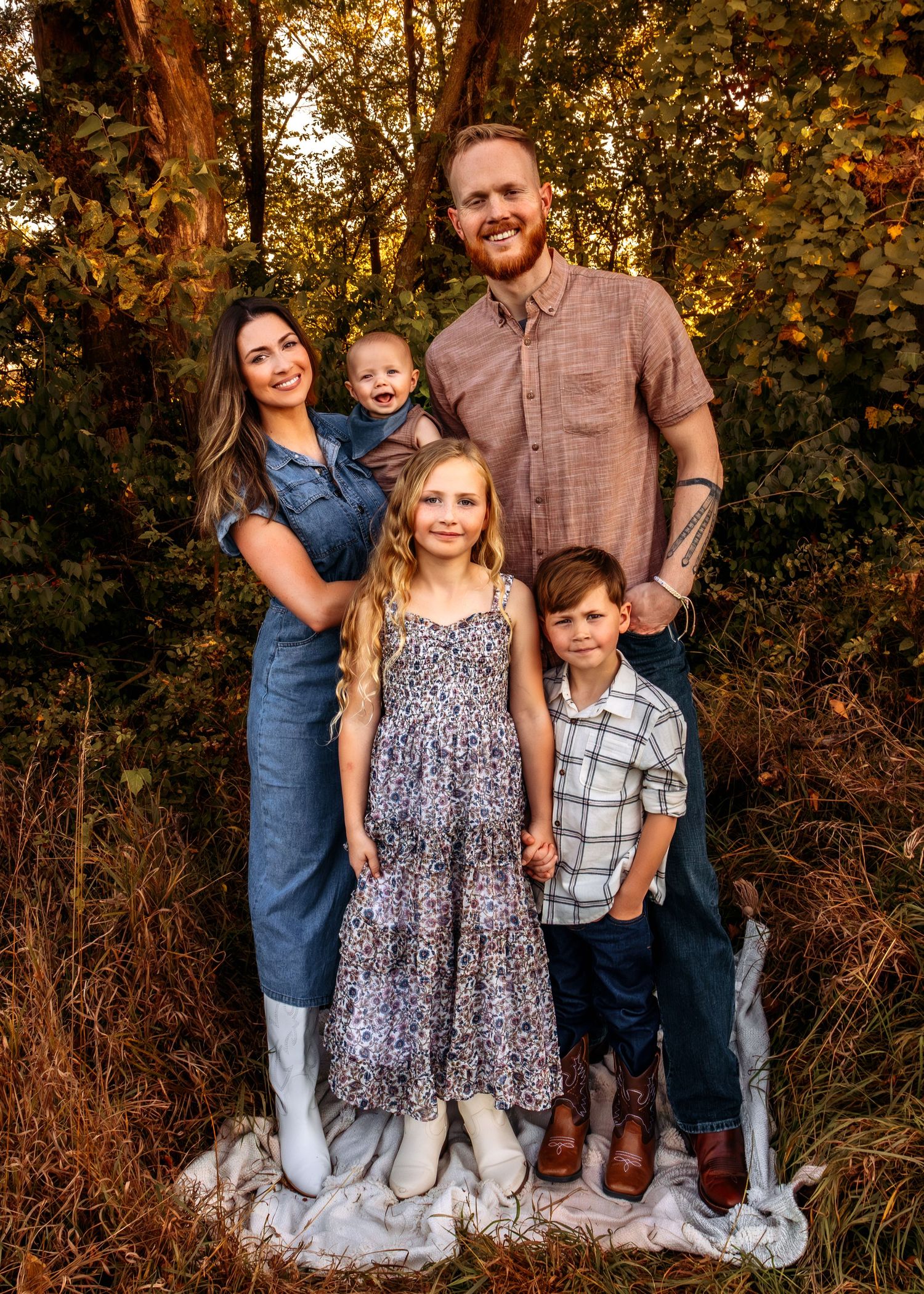 Family of five posing outdoors in golden hour light, parents with three children in coordinated outfits among lush greenery.