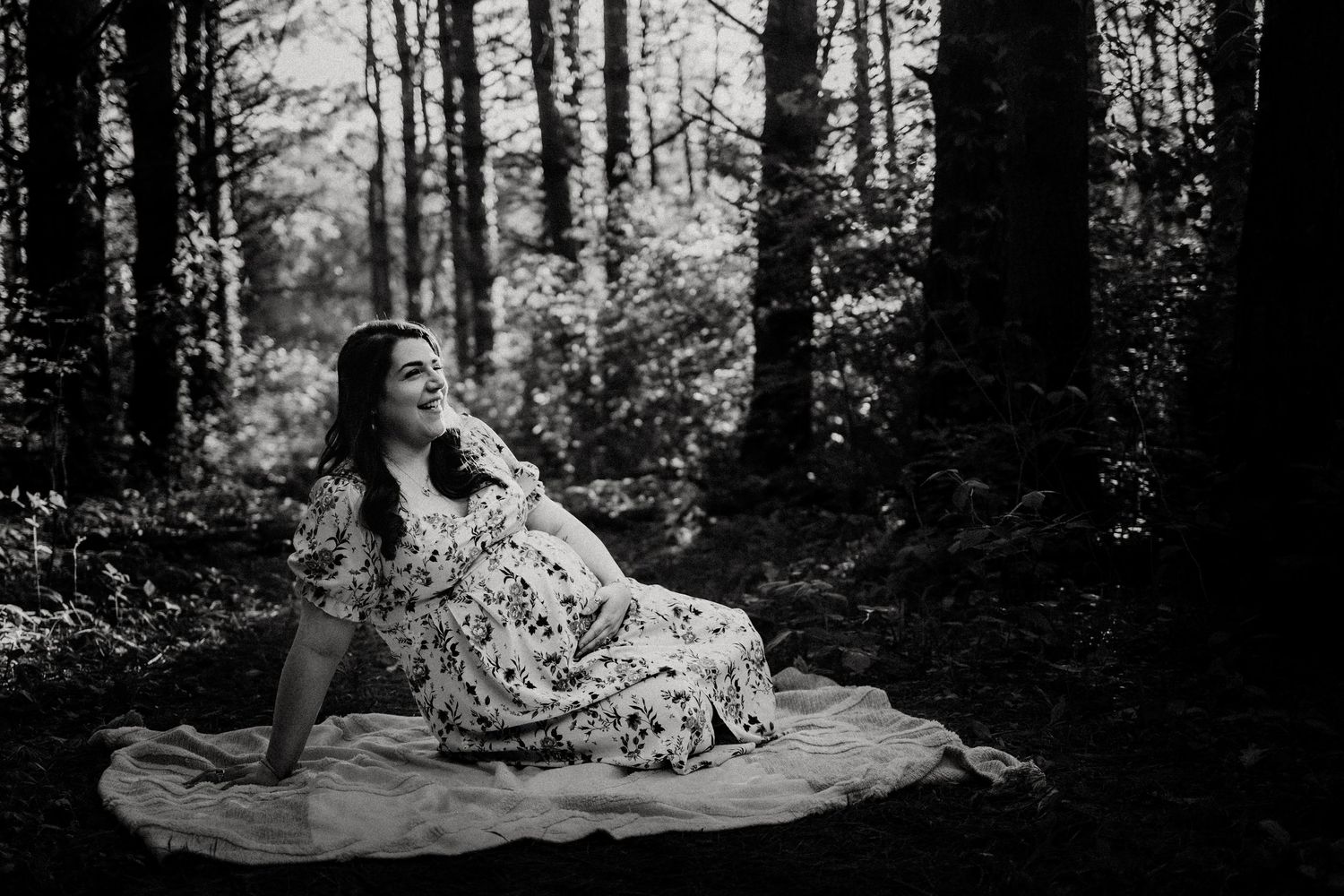 Black and white maternity photo of a pregnant woman in a floral dress sitting peacefully in a forest.