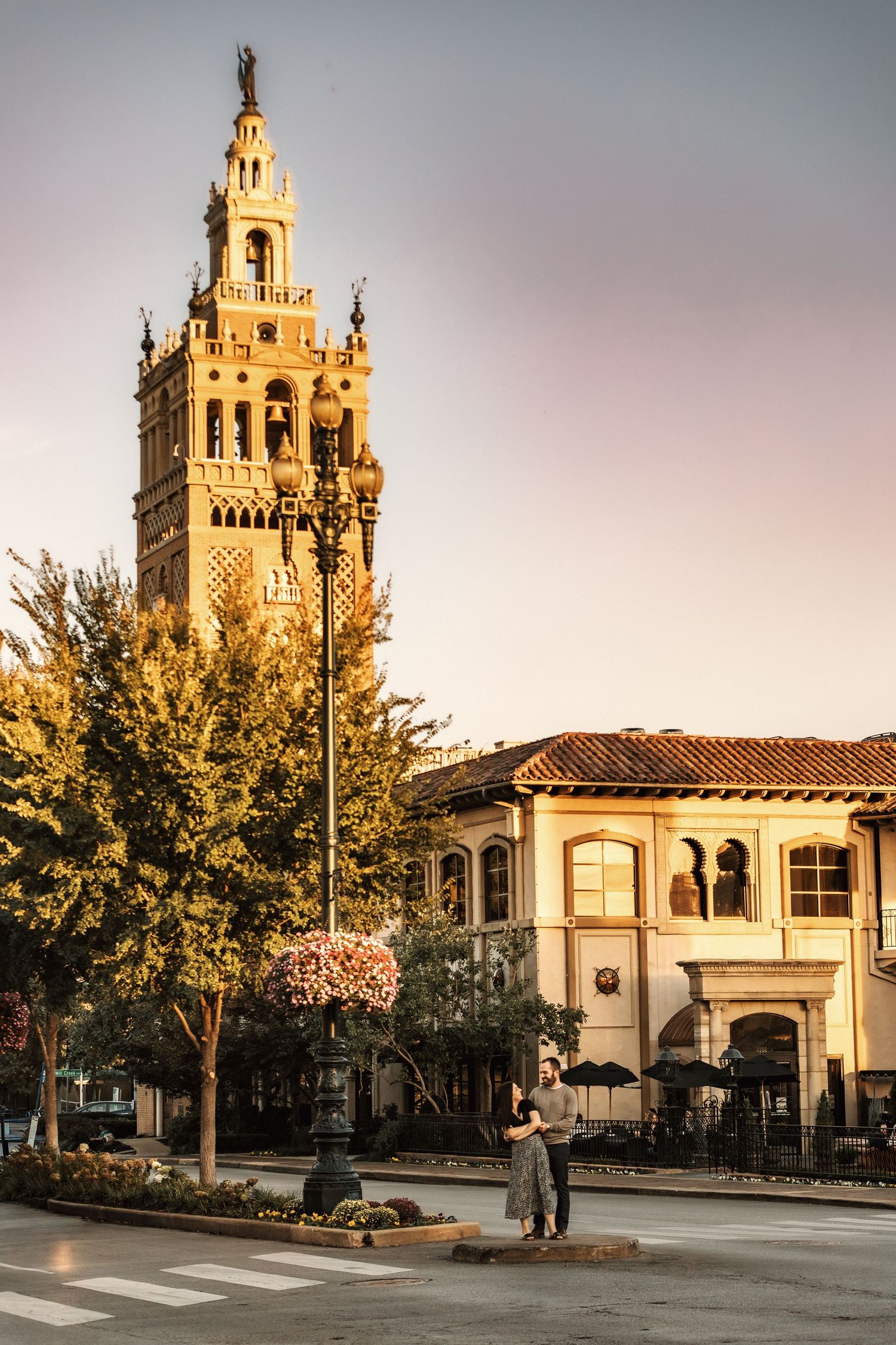 Couple embracing at golden hour near ornate bell tower and Spanish-style building with lush trees.