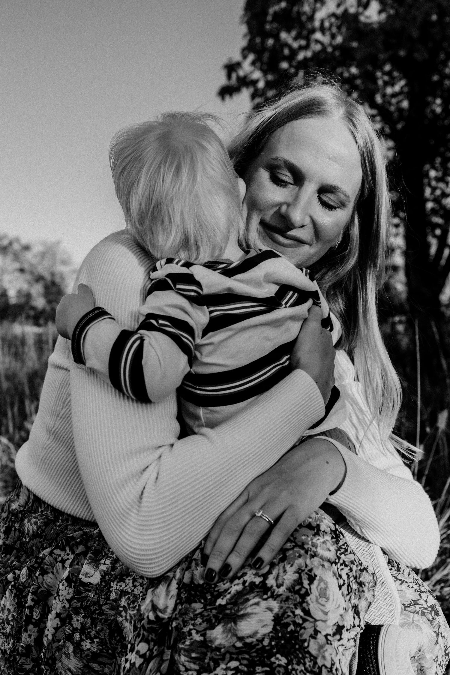 Black and white photo of a smiling mother tenderly embracing her young blonde child outdoors.