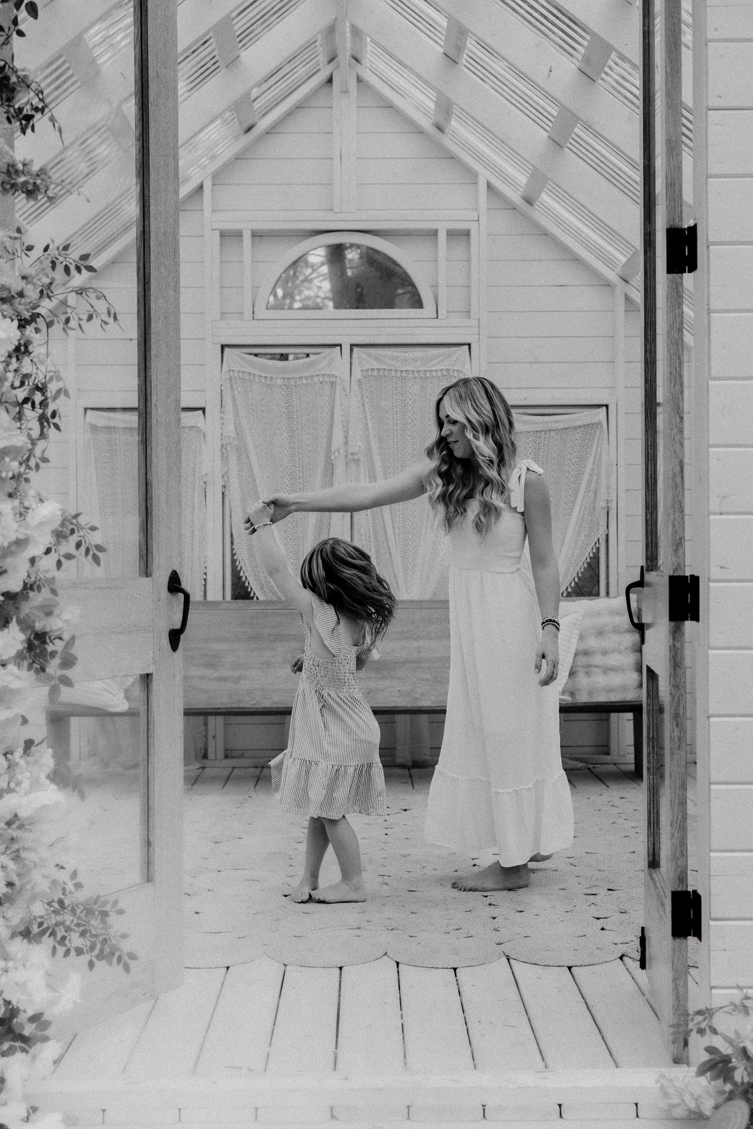 Black and white photo of a woman in a white dress twirling a young girl on a wooden porch of a charming white cottage.