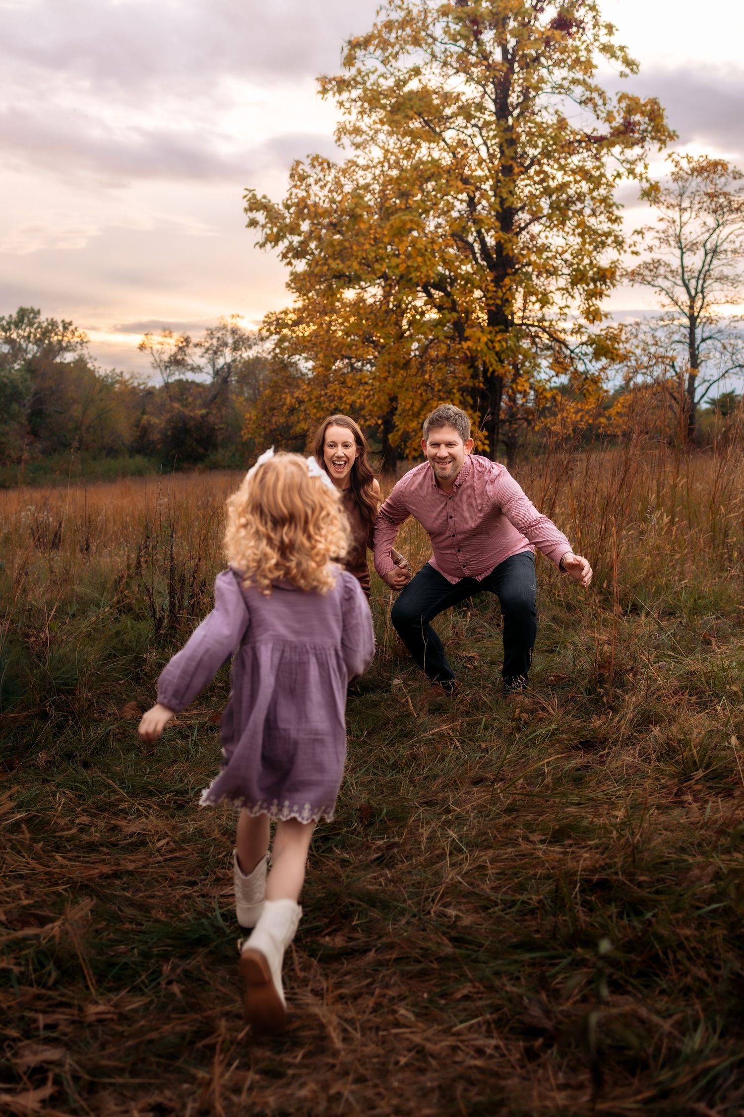 A joyful family moment as a curly-haired girl in purple runs toward her laughing parents in an autumn field.