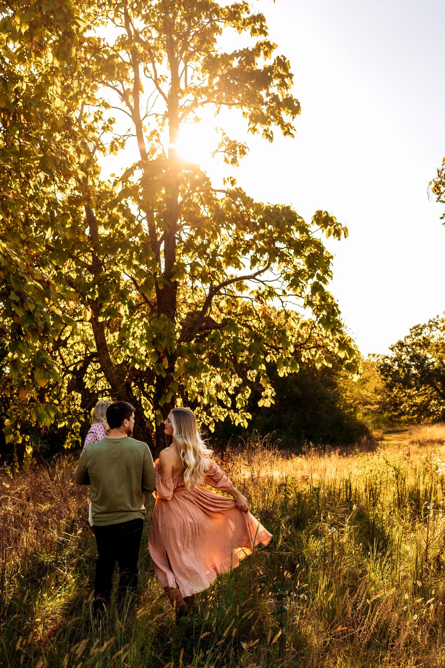 A couple walks through a sunlit meadow toward a large tree, golden hour light streaming through leaves.