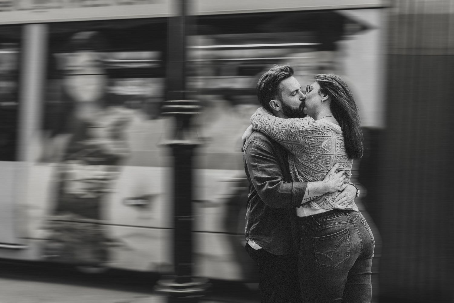 Black and white photo of a couple embracing and kissing in front of a moving tram on a city street.
