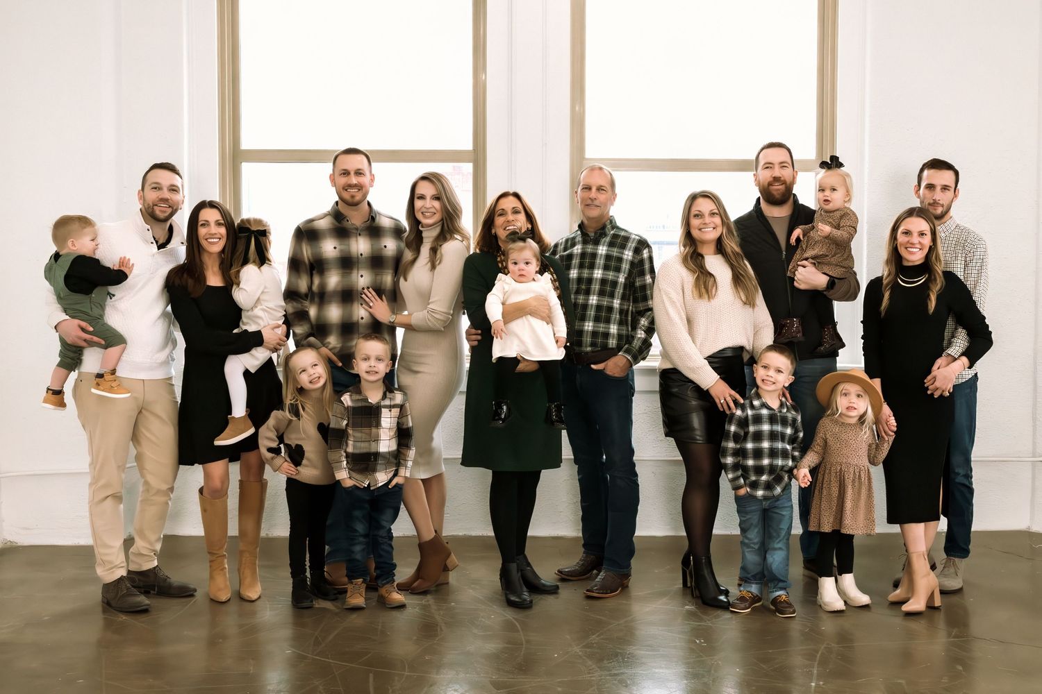 Large family group portrait with adults and children posing together in a bright studio wearing coordinated neutral and plaid outfits.