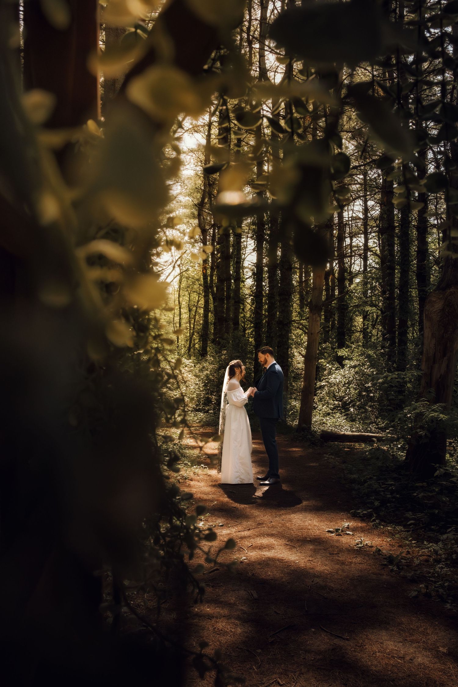 A bride and groom share a romantic moment on a forest path, framed by lush green leaves and tall trees.