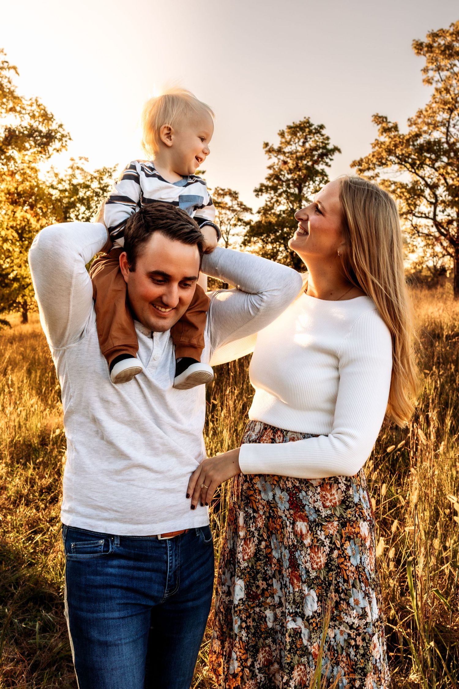 Happy young family with toddler on dad's shoulders enjoying golden hour sunset in a field.