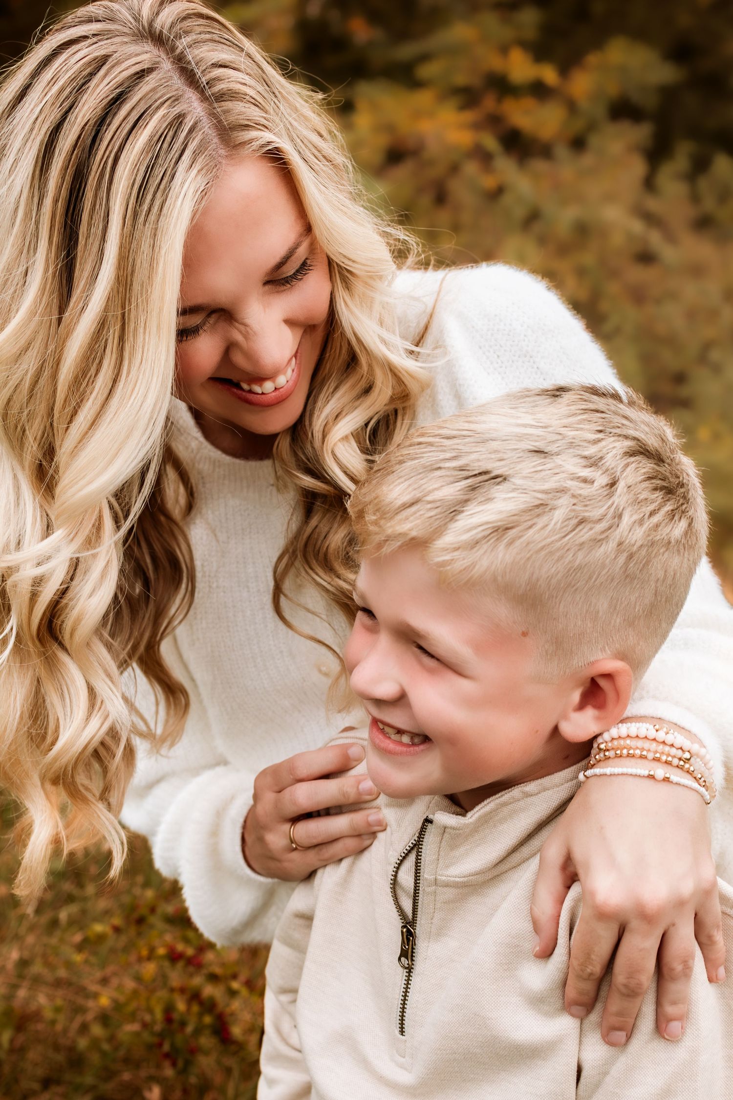 A smiling blonde woman hugs a laughing young blonde boy outdoors in autumn, both wearing cream sweaters.