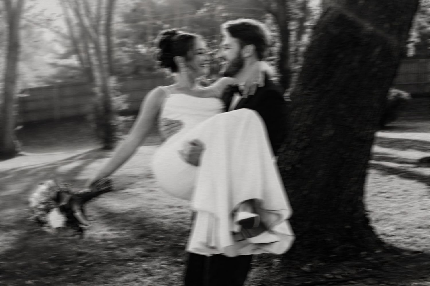 Groom carrying bride in white dress outdoors in black and white wedding photo near trees.