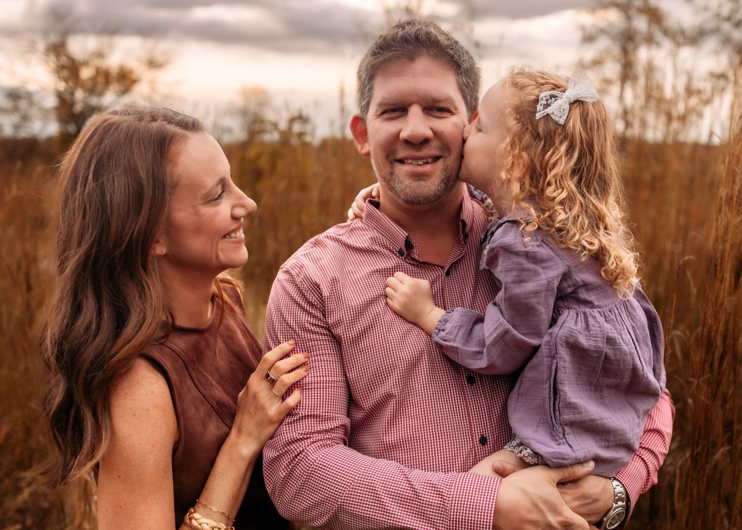Happy family portrait with parents and young daughter in autumn field with warm golden tones.