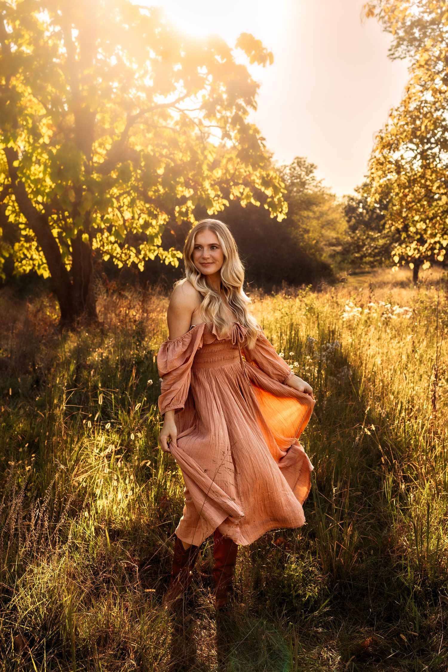 Blonde woman in flowing peach off-shoulder dress and brown boots standing in sunlit meadow with golden trees.