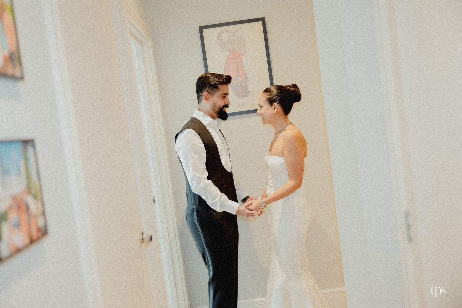 A bride and groom share a tender first look moment, holding hands in a bright hallway on their wedding day.