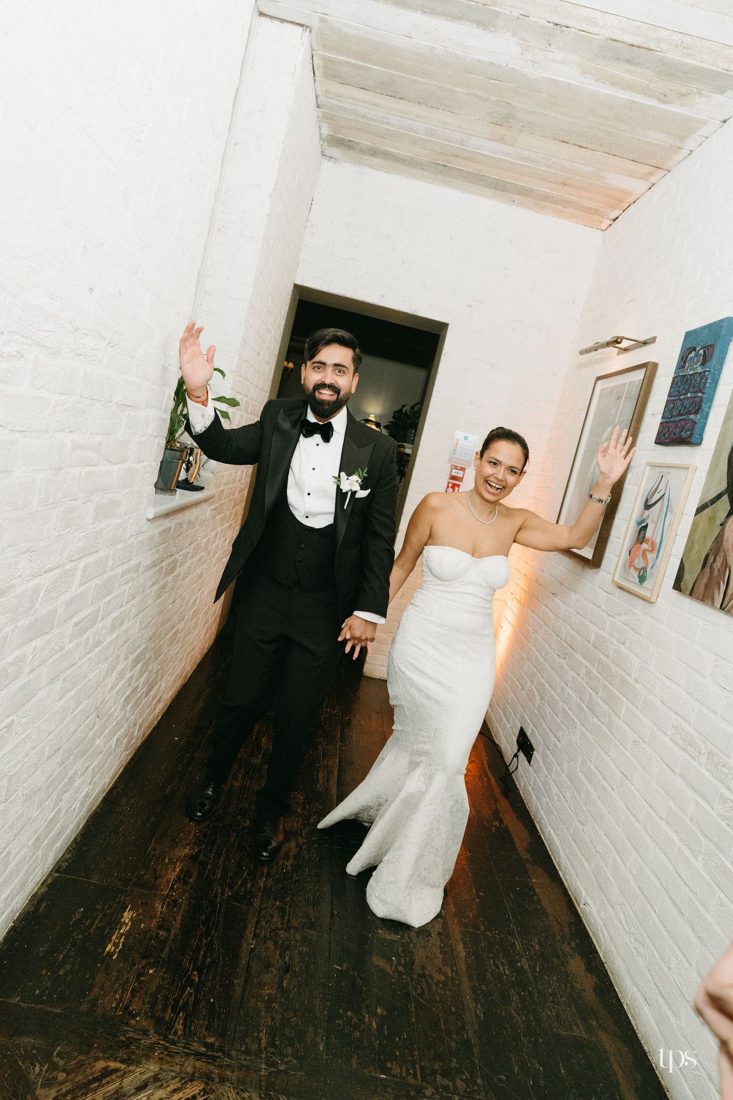 Bride and groom in wedding attire wave joyfully while walking through a narrow white brick hallway.