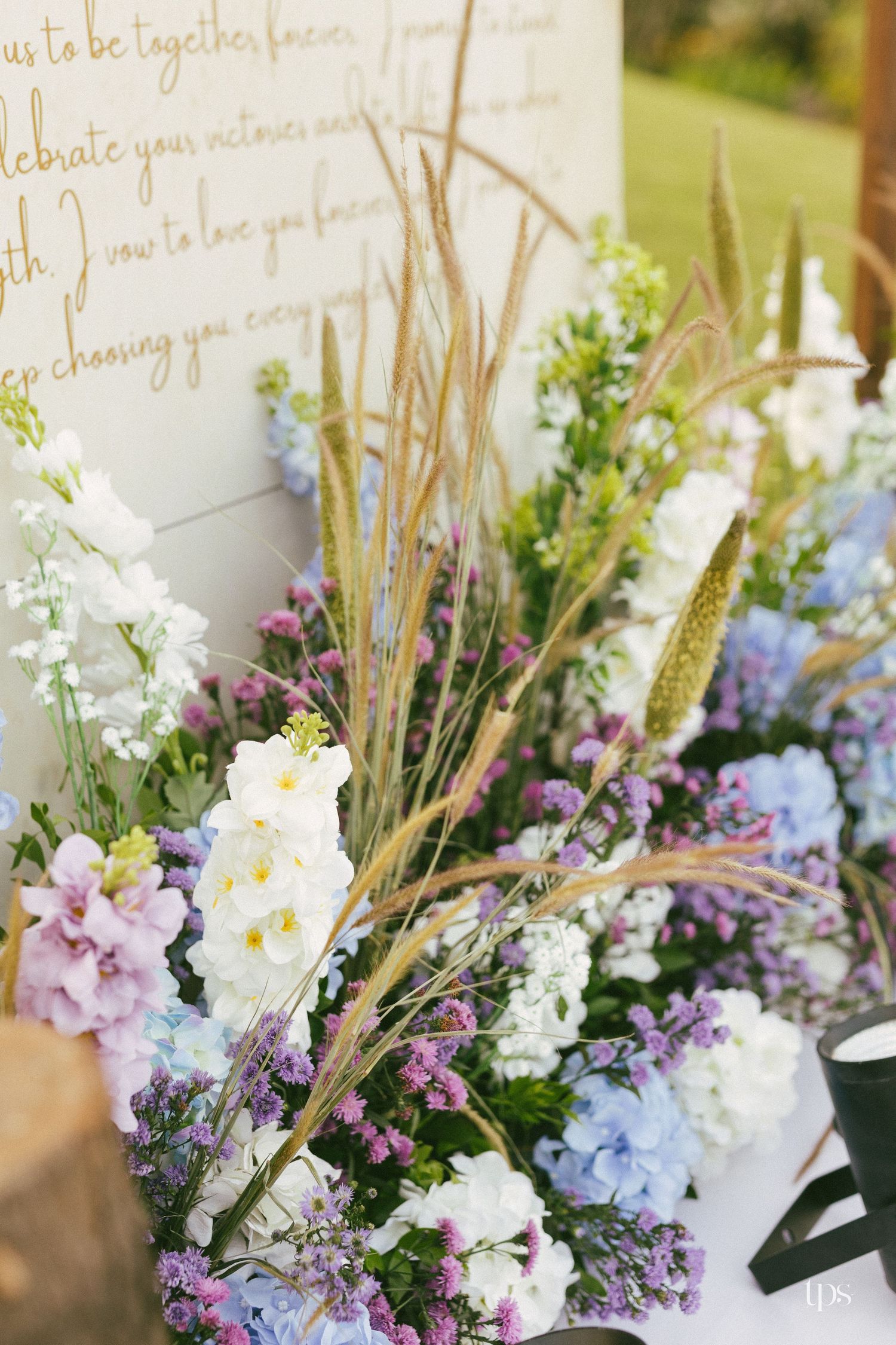 Wedding ceremony floral arrangement of white delphinium, purple blooms and tall wheat grasses beside a sign with handwritten vows.