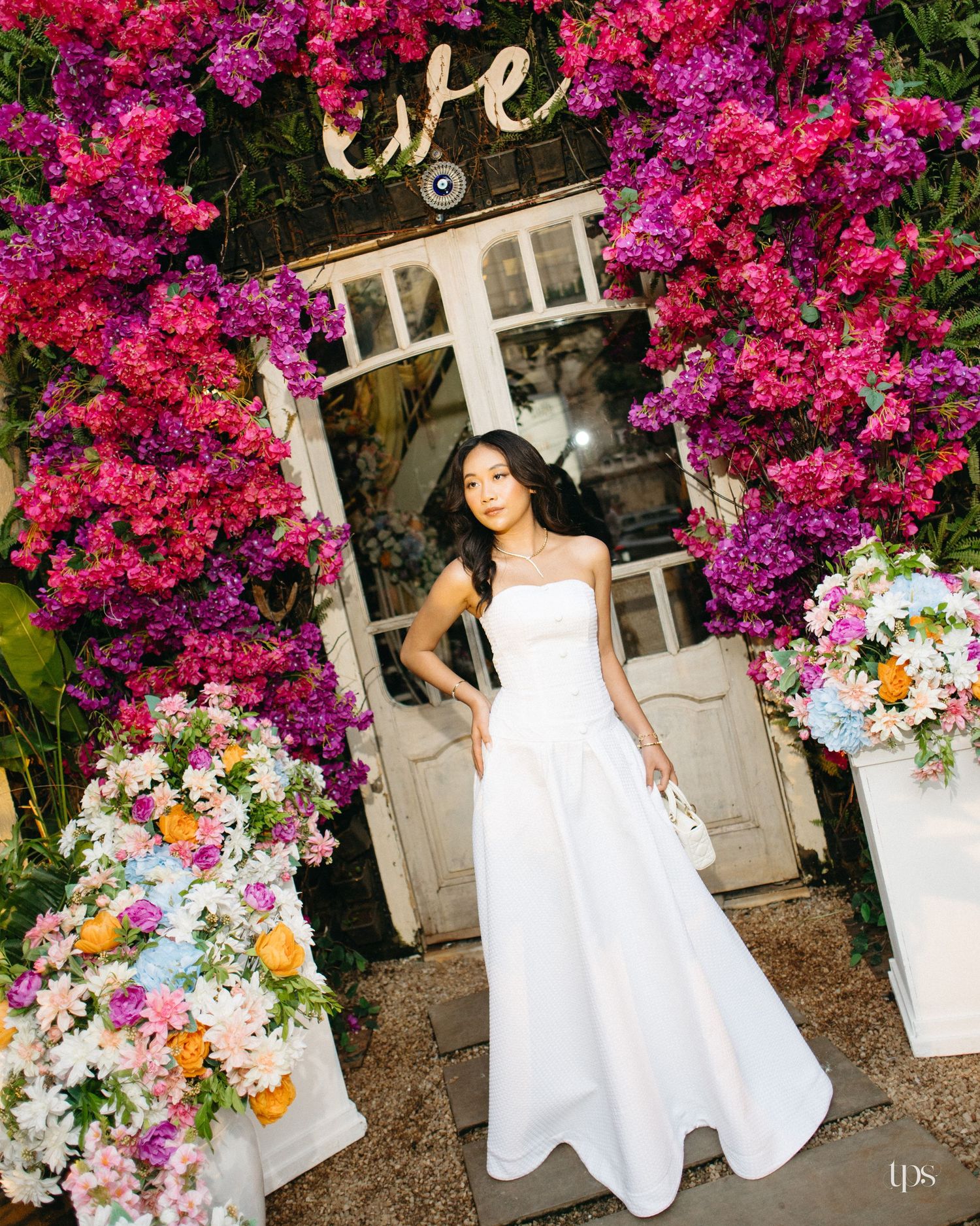 Woman in a white strapless gown stands before a white door framed by vibrant pink bougainvillea and floral arrangements.