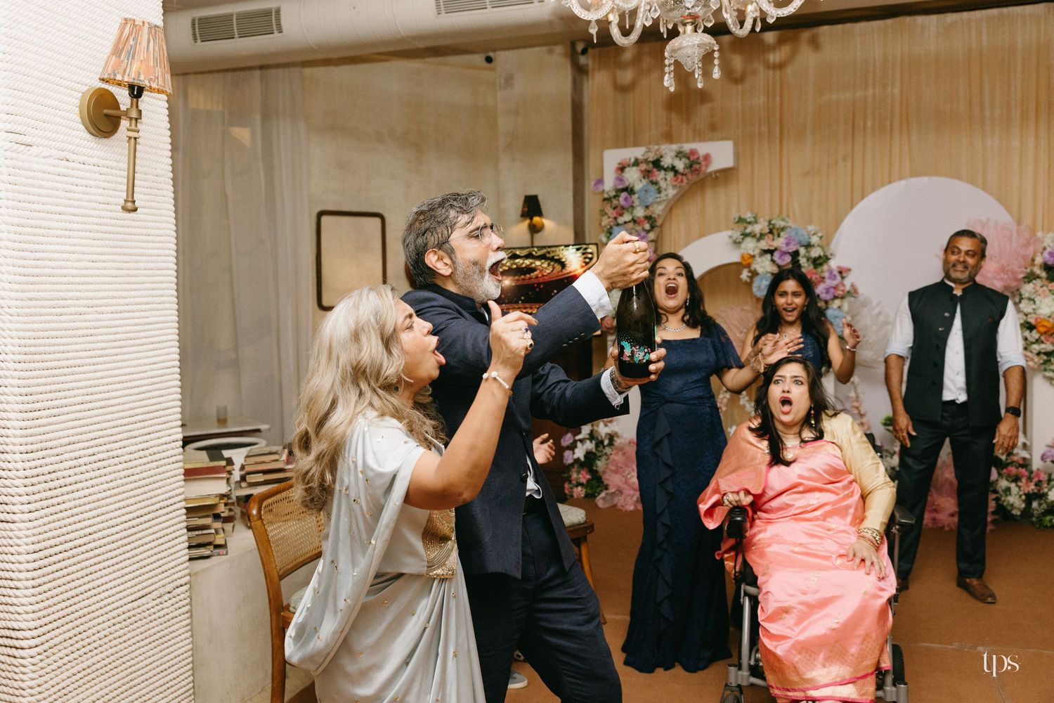 Guests celebrate at an elegant indoor party, raising glasses and toasting around a woman in a pink dress seated in a wheelchair.
