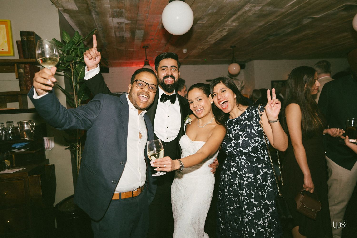 Wedding party guests celebrating with drinks, bride in white gown and groom in tuxedo posing joyfully indoors.