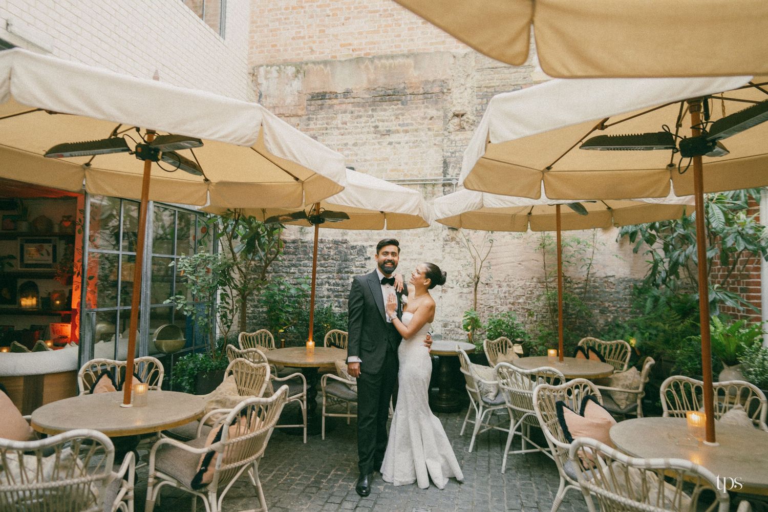 Bride and groom pose together on a romantic outdoor patio with wicker chairs, candles, and cream umbrellas at their wedding.