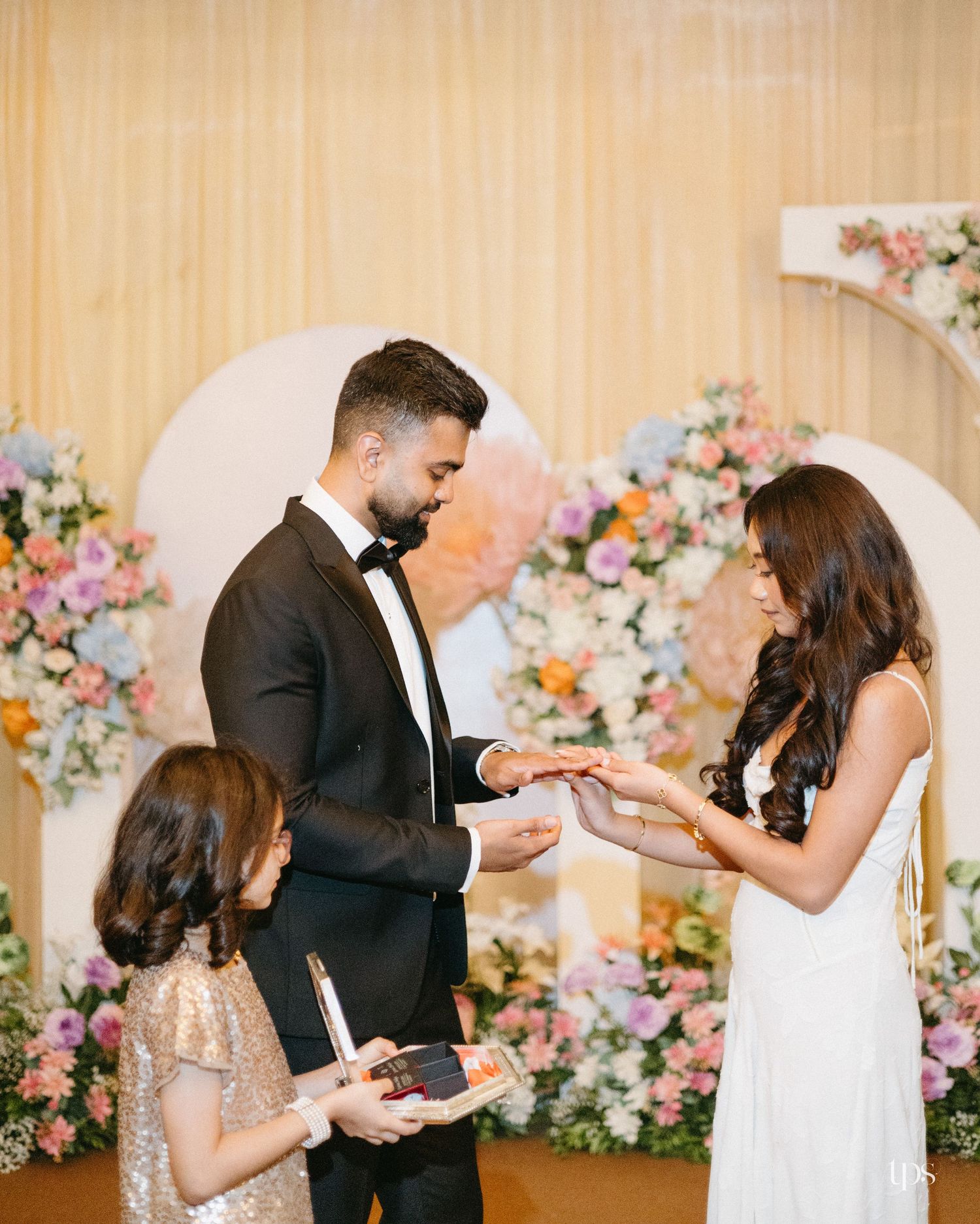 Groom in a black tuxedo and bride in a white dress exchange rings at a floral wedding ceremony while a young girl holds the ring box.