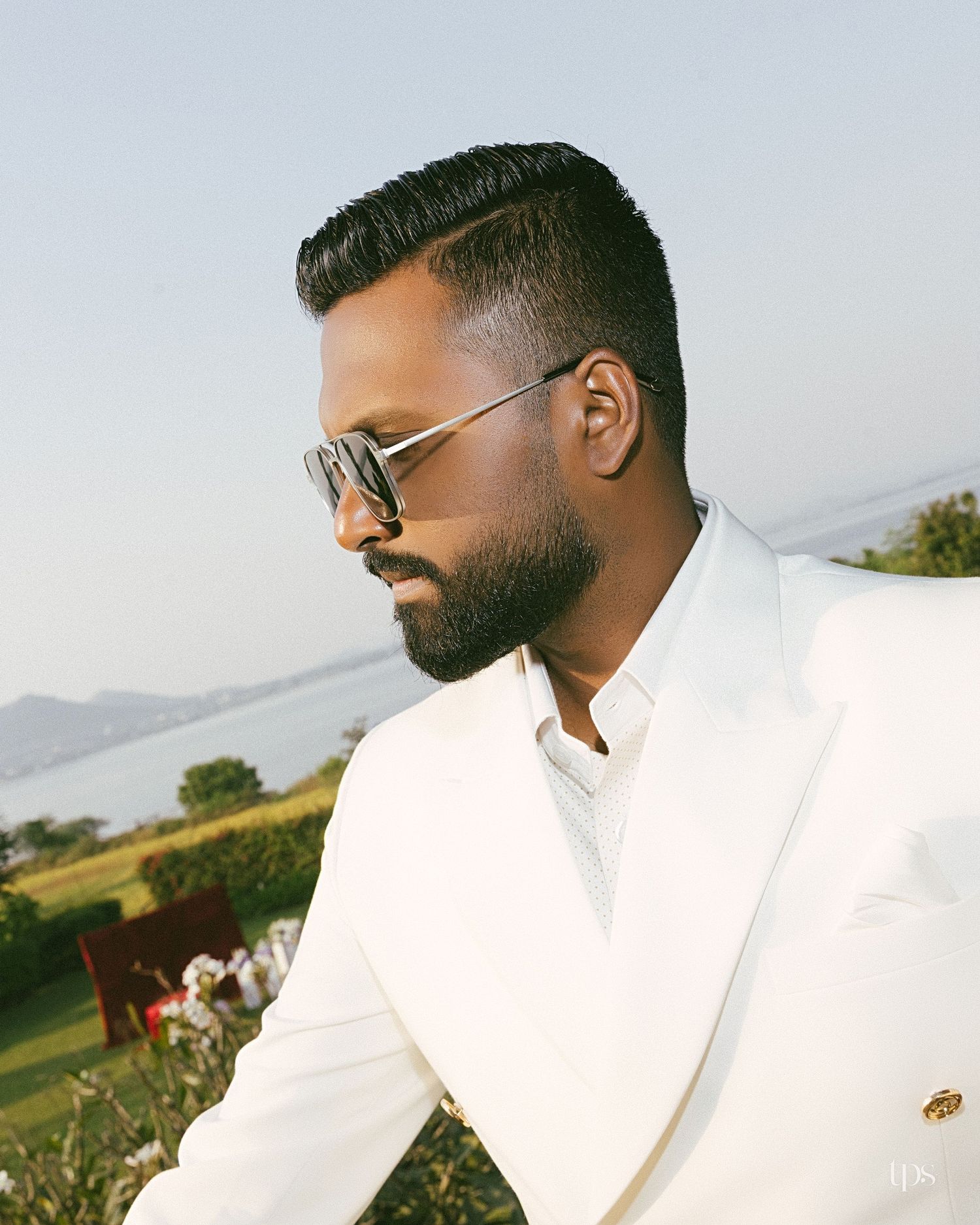 Bearded man in sunglasses and a white double-breasted blazer stands in profile at an outdoor event with a scenic lake and hills behind him.