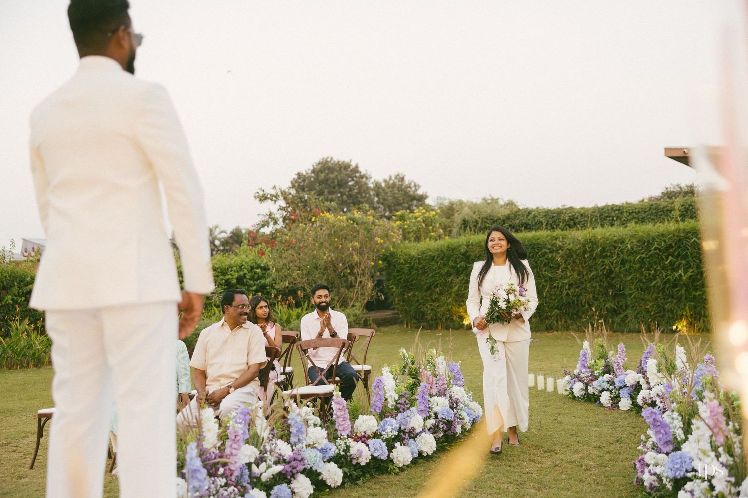 Bride in white carrying a bouquet walks down an outdoor garden aisle lined with purple and white flowers as the groom and guests watch.