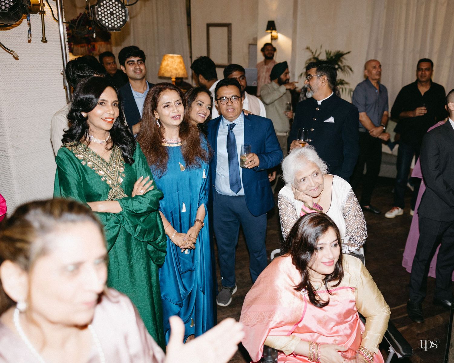 Guests in elegant formal attire, including women in green and blue dresses and a man in a suit, mingle at a festive indoor celebration.