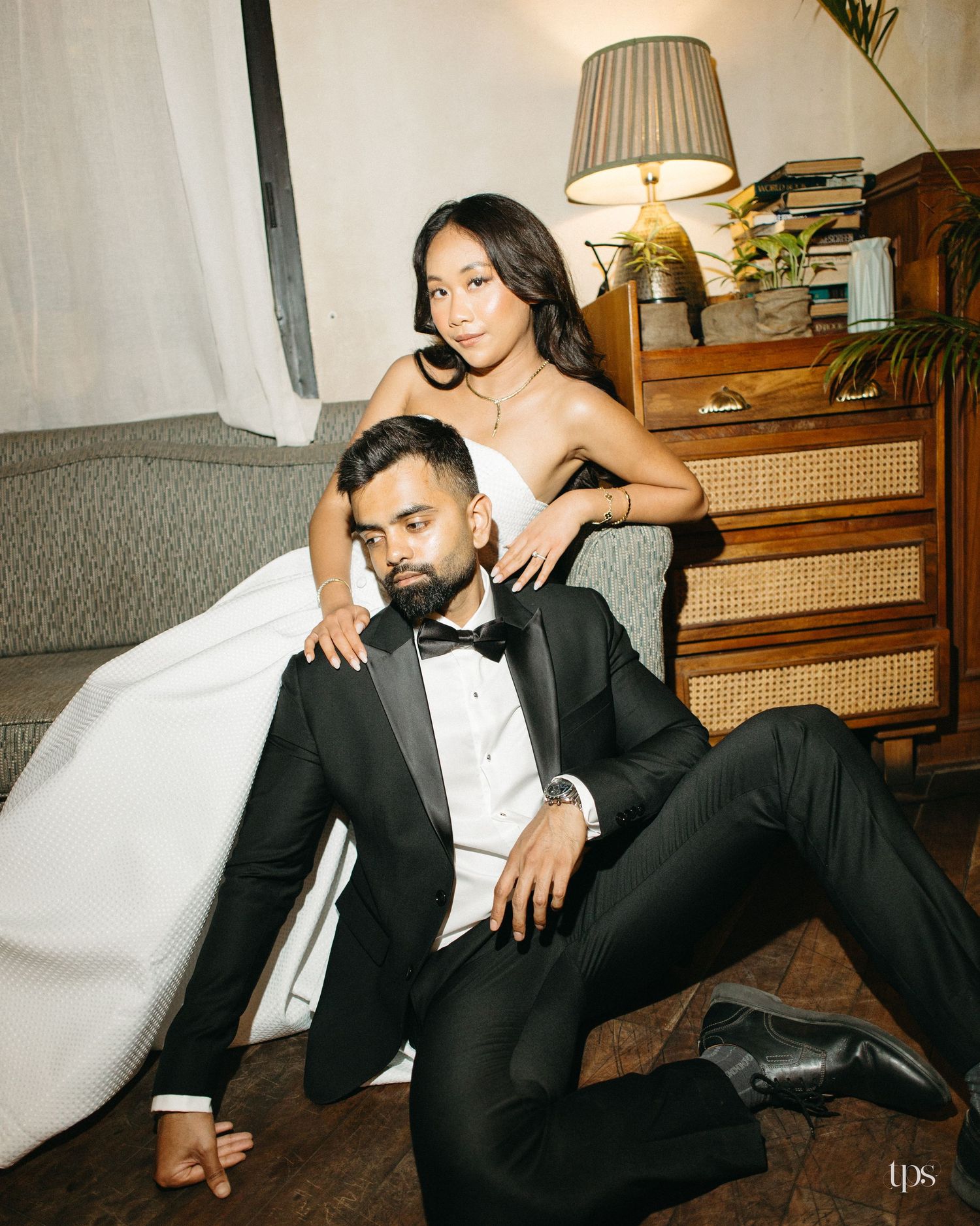 Bride in a white strapless gown leans over a groom in a black tuxedo and bow tie seated on a wood floor in a cozy vintage room.