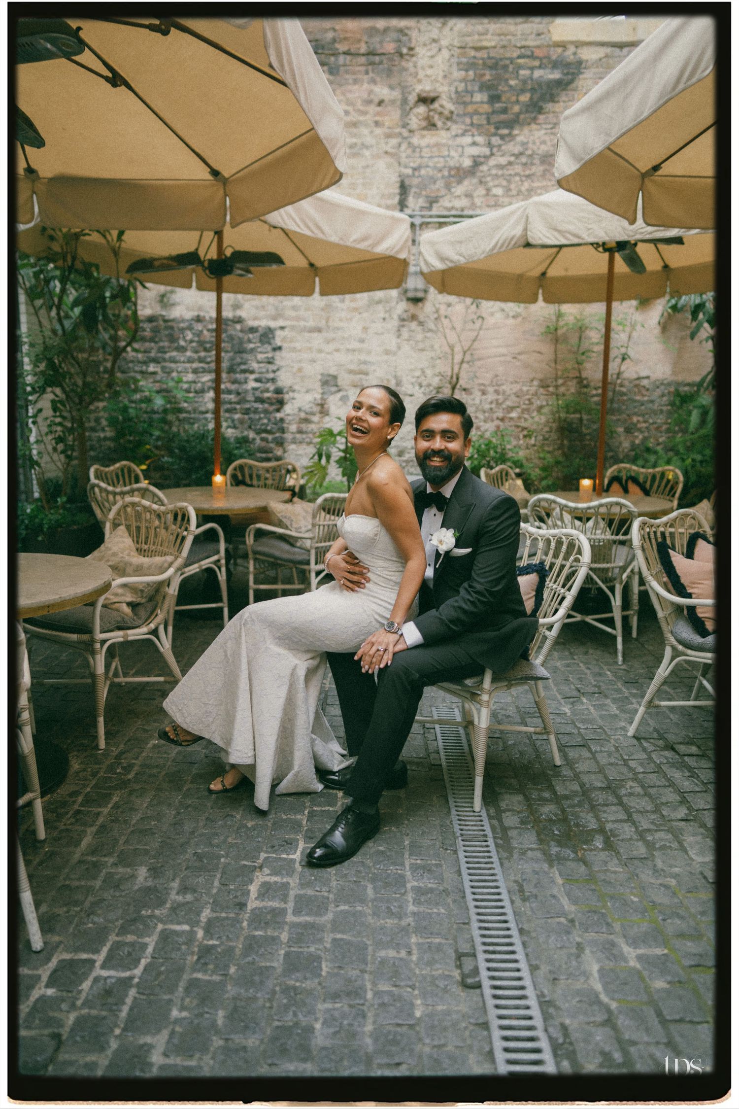 Bride and groom sitting together in a charming outdoor courtyard with rattan chairs and market umbrellas on their wedding day.